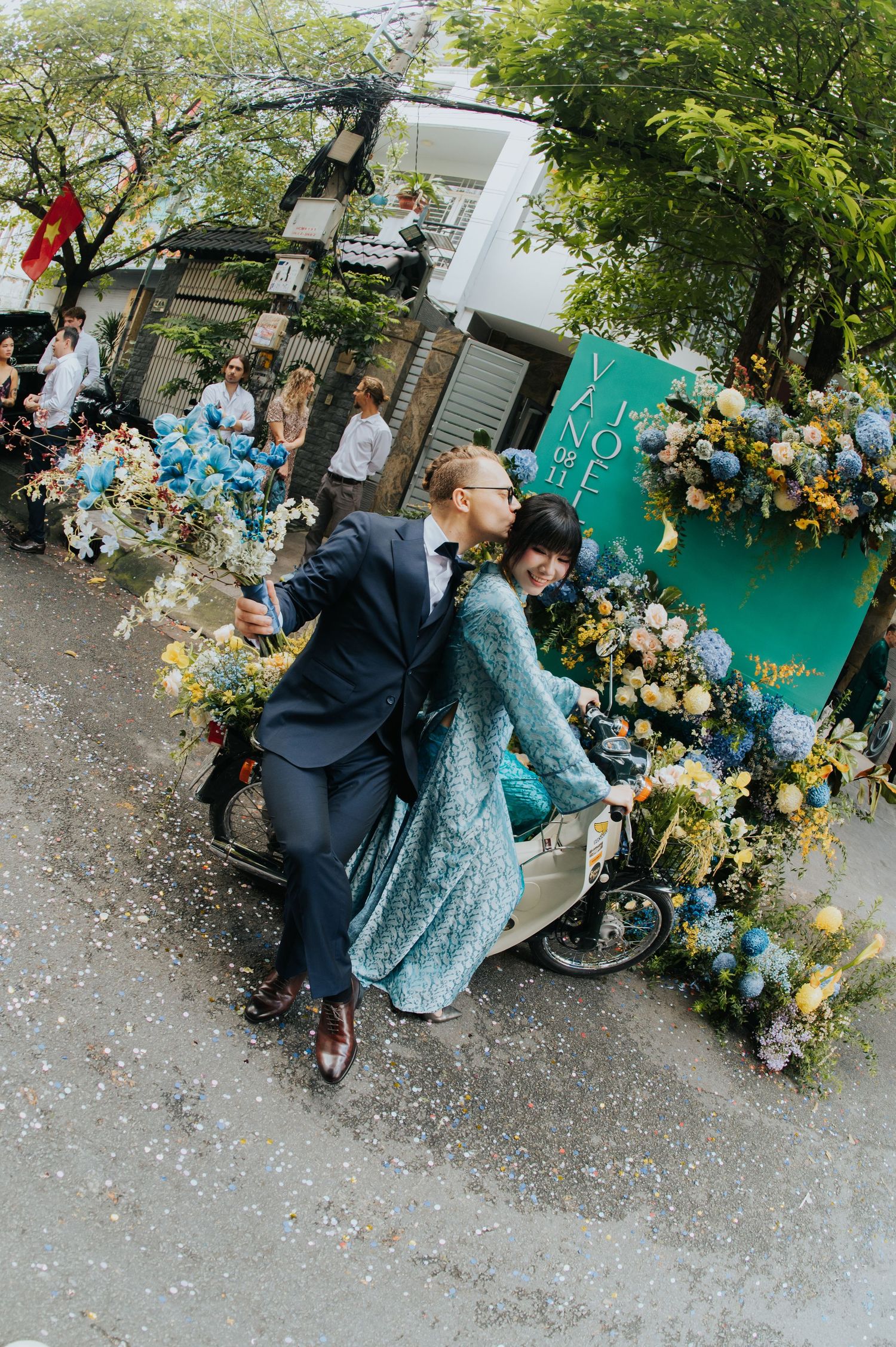 Couple in blue outfits riding a flower-adorned scooter on a street, surrounded by lush floral arrangements.
