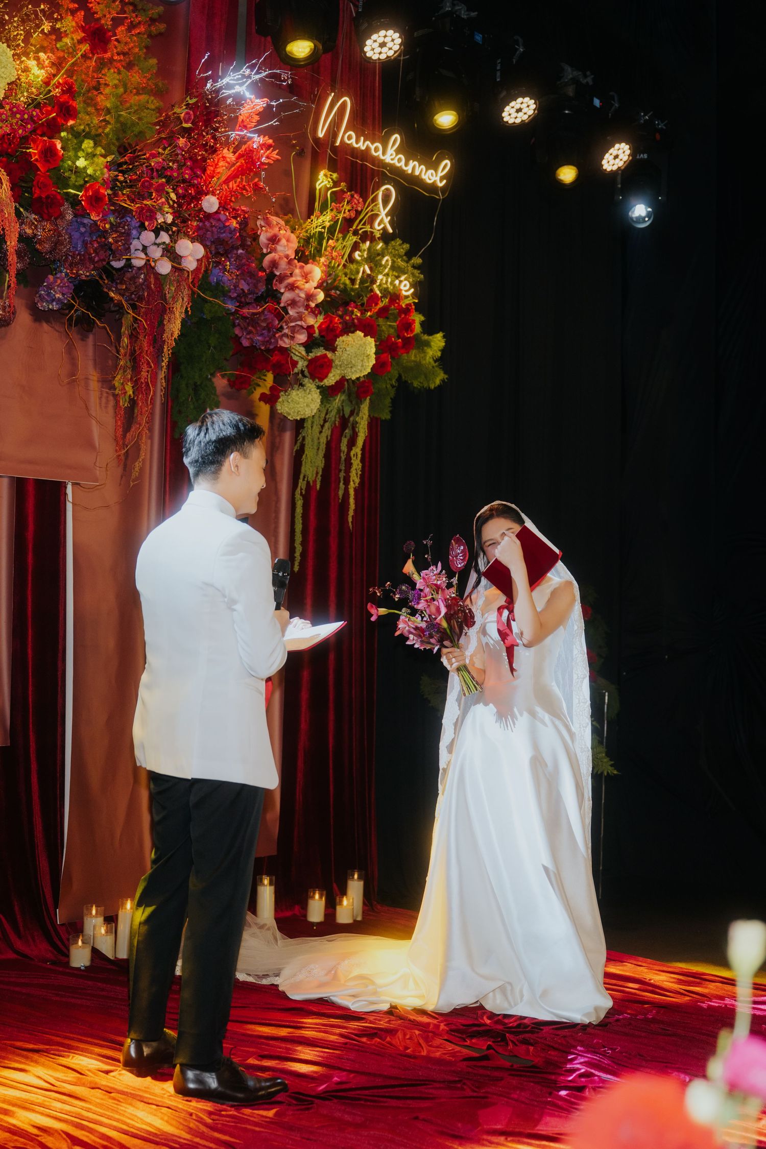 Bride and groom exchange vows at elegant wedding ceremony with lush floral backdrop and romantic neon sign lighting.