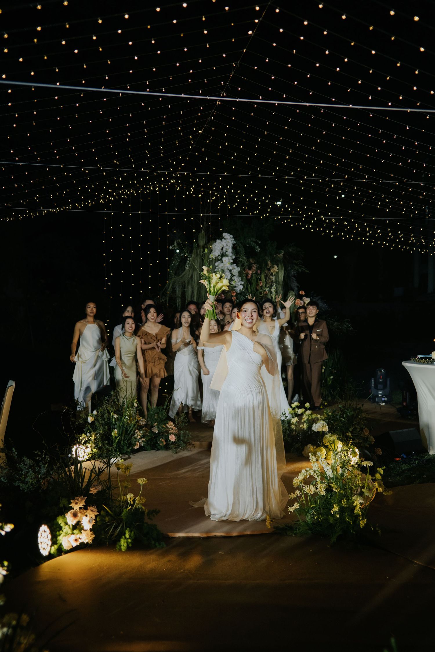 Bride tossing bouquet at elegant outdoor wedding reception under twinkling star lights with guests gathered behind.
