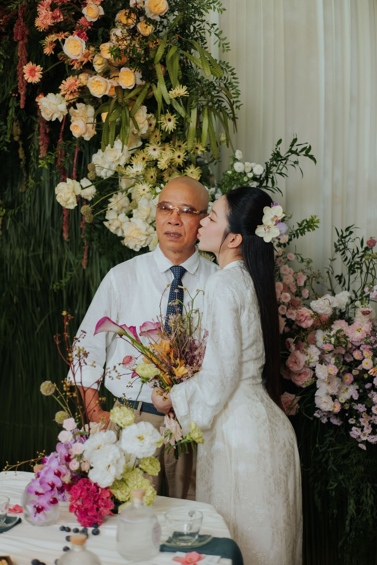 Bride in white dress kissing elderly man on cheek at floral wedding ceremony backdrop with colorful blooms.