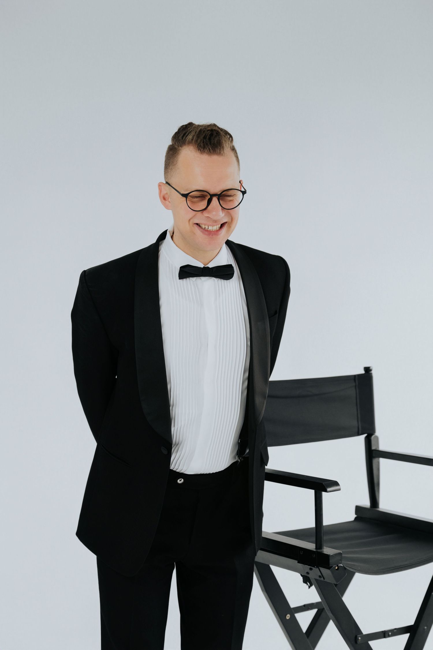 Smiling man in black tuxedo and bow tie with glasses stands beside a director's chair against white background.