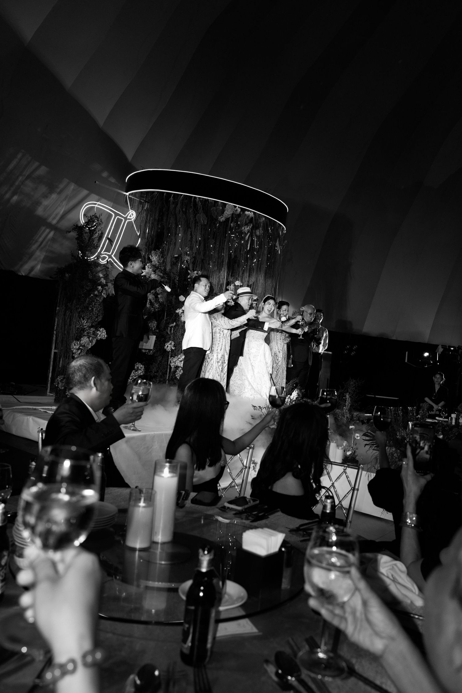 Black and white wedding reception photo showing couple dancing on stage surrounded by guests raising glasses in celebration.