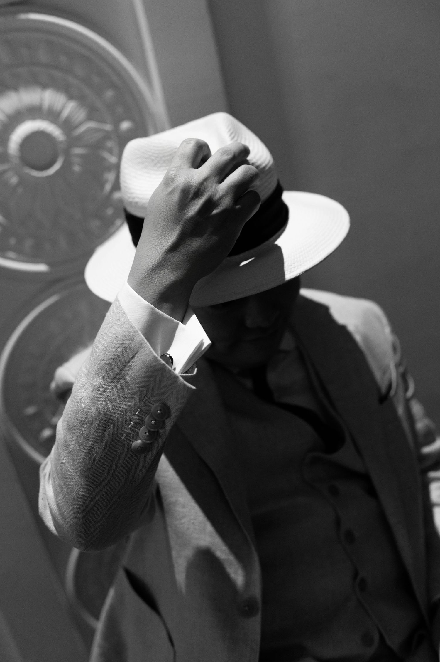 Black and white photo of a man in a suit adjusting his white fedora hat, covering his face dramatically.