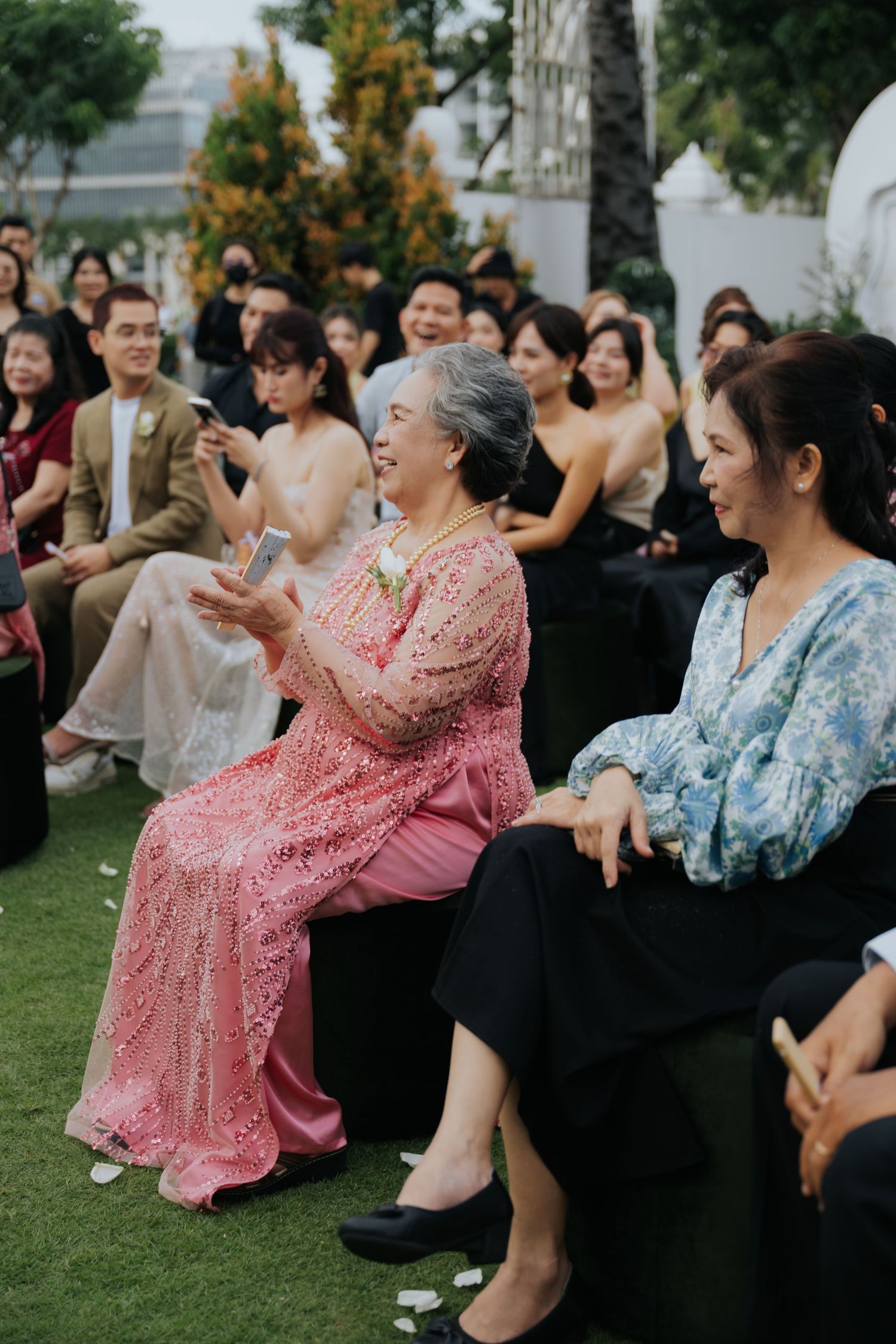 Elderly woman in pink lace dress smiles at outdoor wedding ceremony seated among guests.