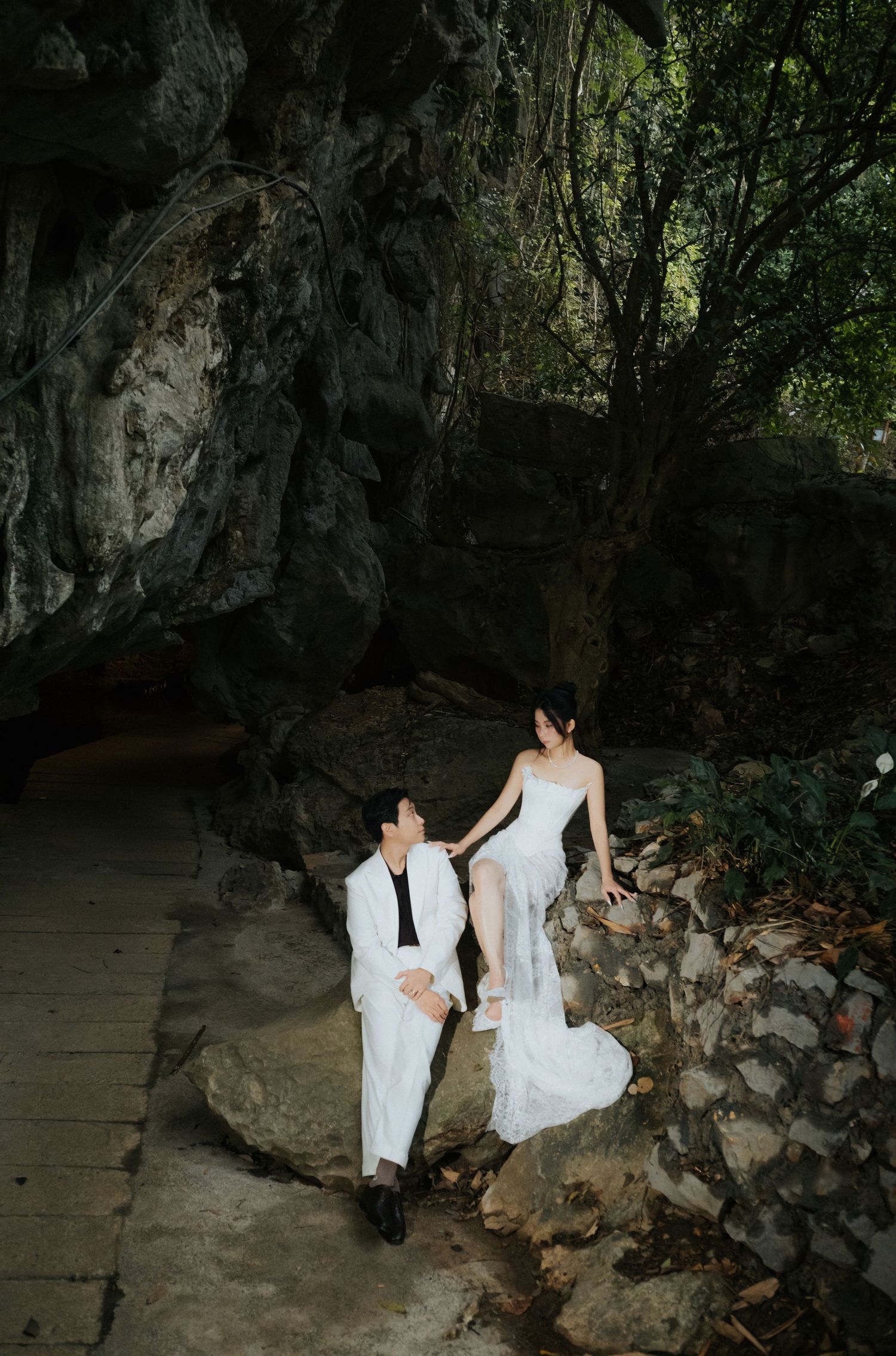 Couple in white wedding attire share a romantic moment on rocks near a dramatic cave entrance.