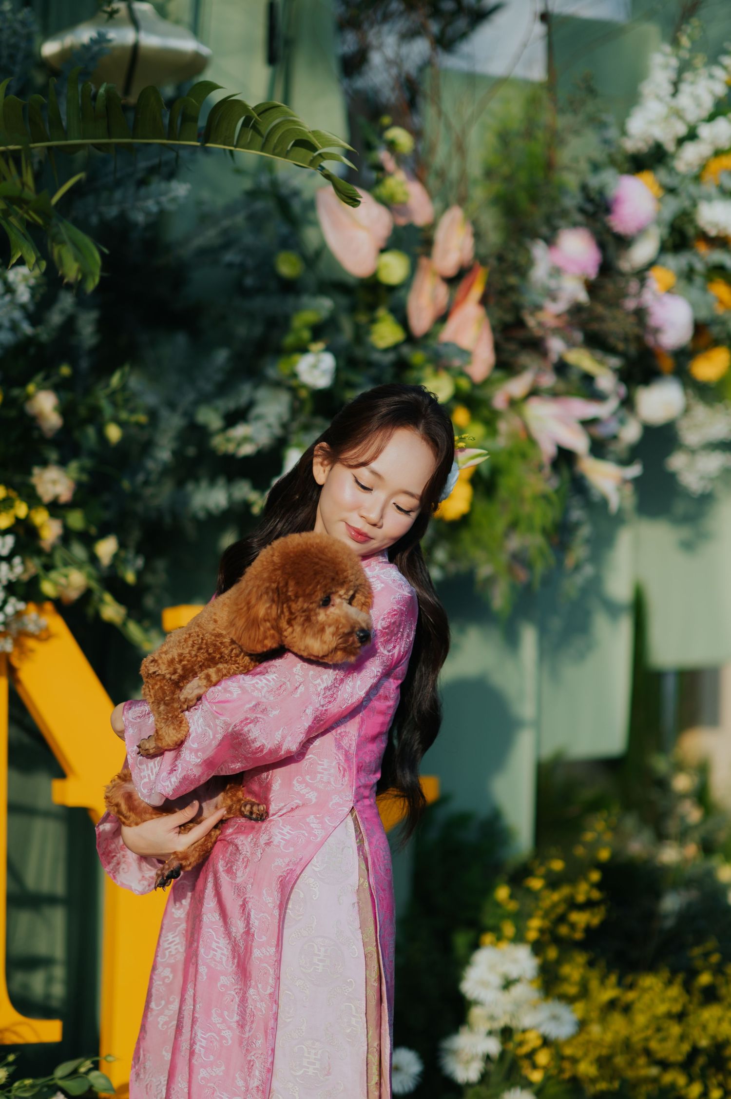 Young woman in pink floral dress tenderly hugging a brown toy poodle puppy surrounded by colorful flowers.