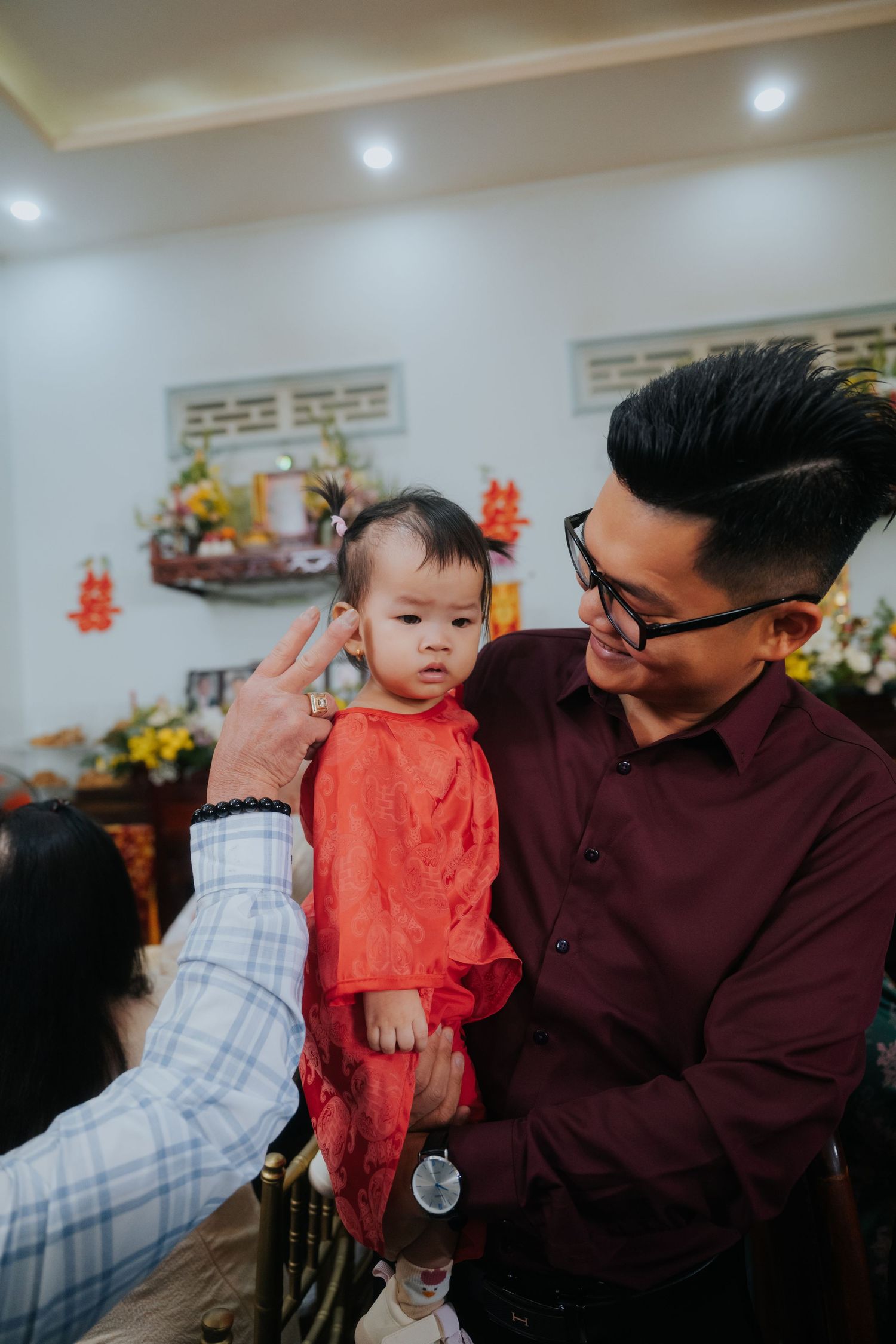 Man in maroon shirt holding baby in red outfit at Asian celebration with altar in background.