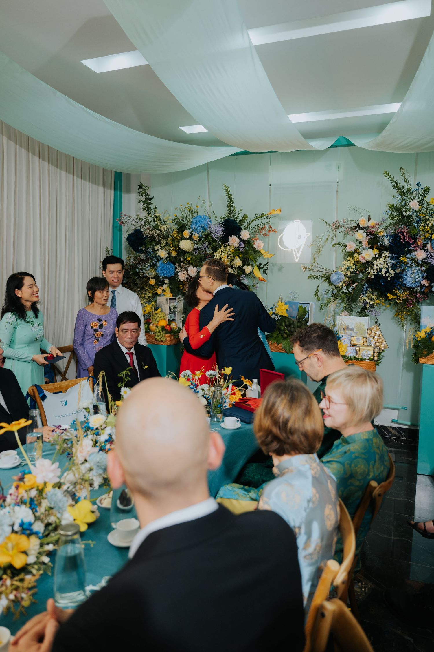 Guests gather at a decorated wedding reception table as the couple shares a kiss near floral arrangements.