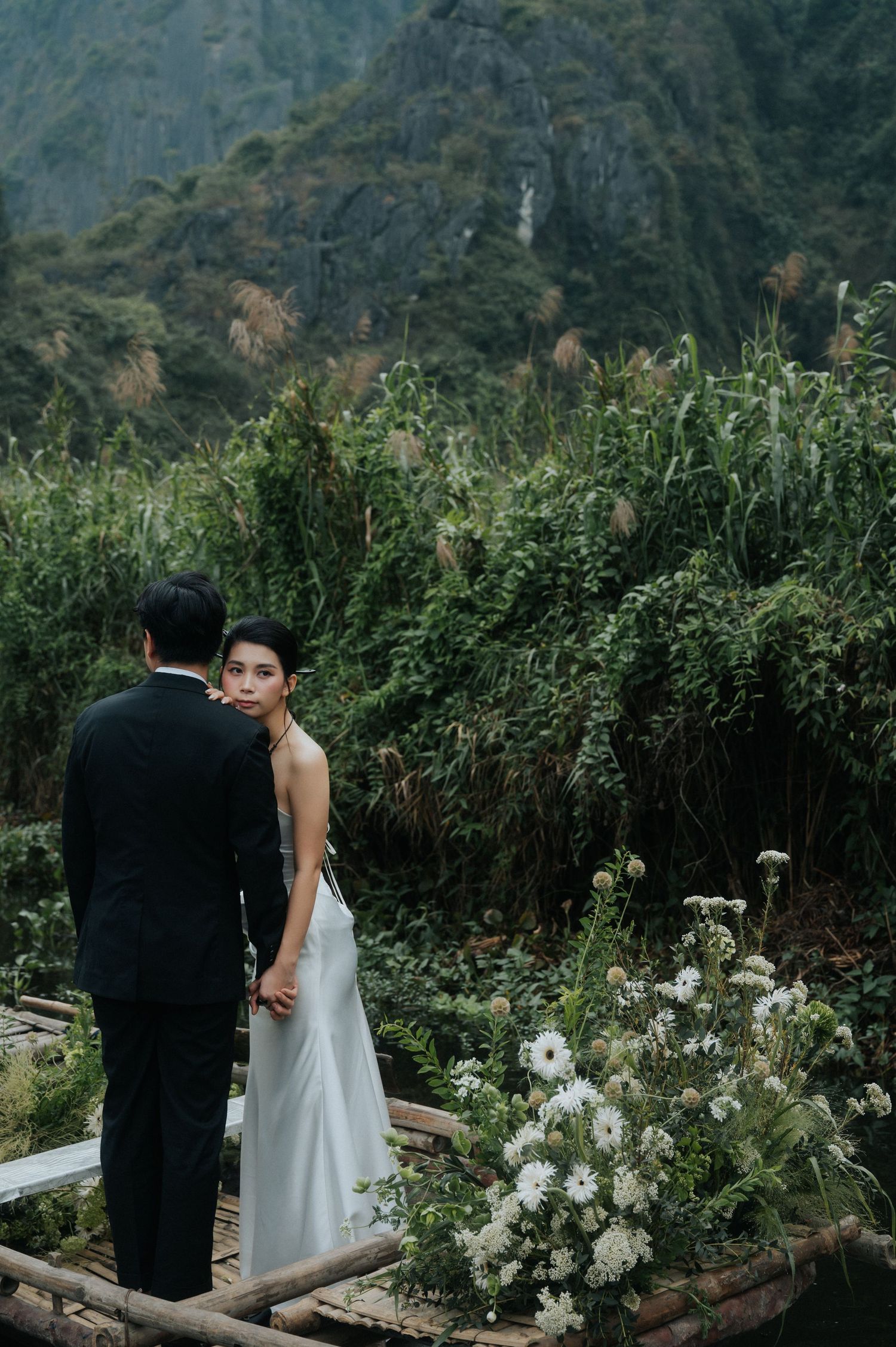 Couple in wedding attire standing before lush green mountains, bride in white gown glancing back at camera.