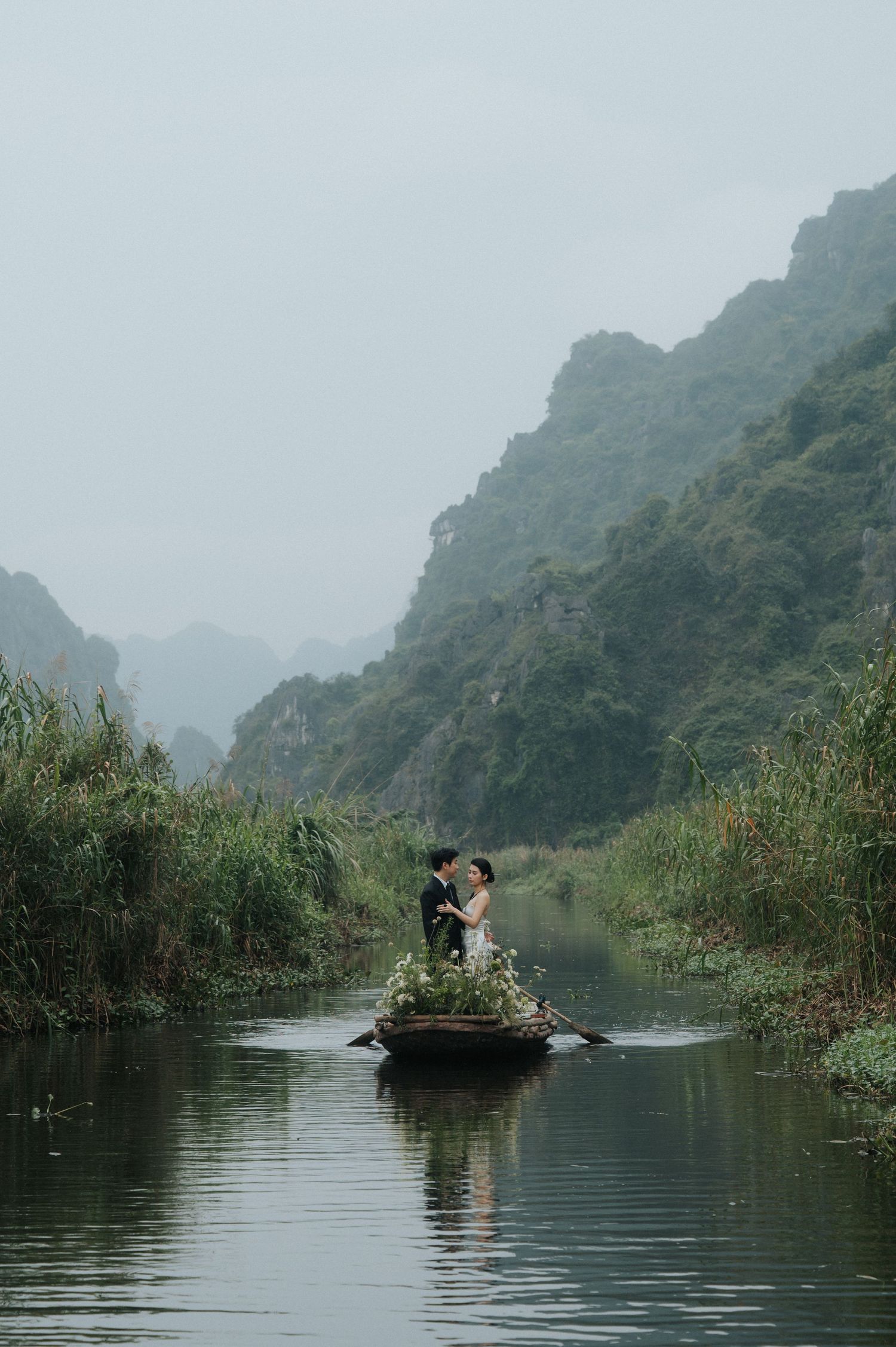 A couple stands on a flower-adorned boat on a misty river surrounded by lush greenery and karst mountains.