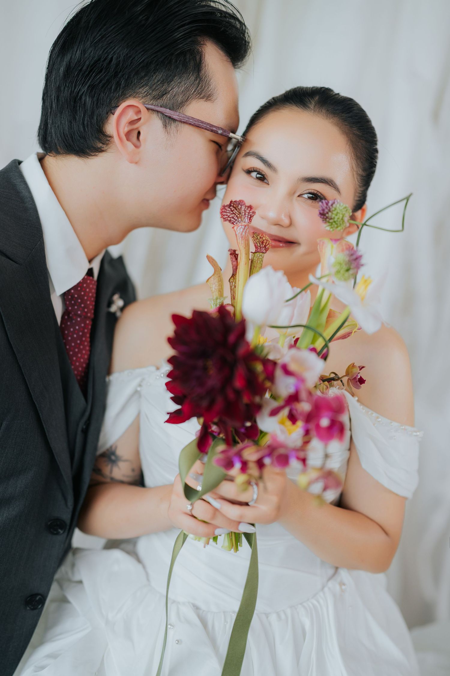A groom in a gray suit kisses his bride on the cheek as she smiles holding a vibrant floral bouquet.