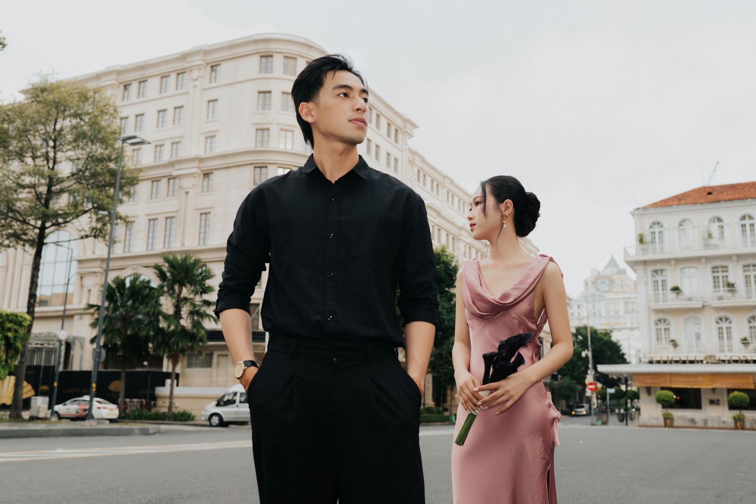 A stylish young Asian couple poses on a city street, man in black shirt and woman in pink dress.
