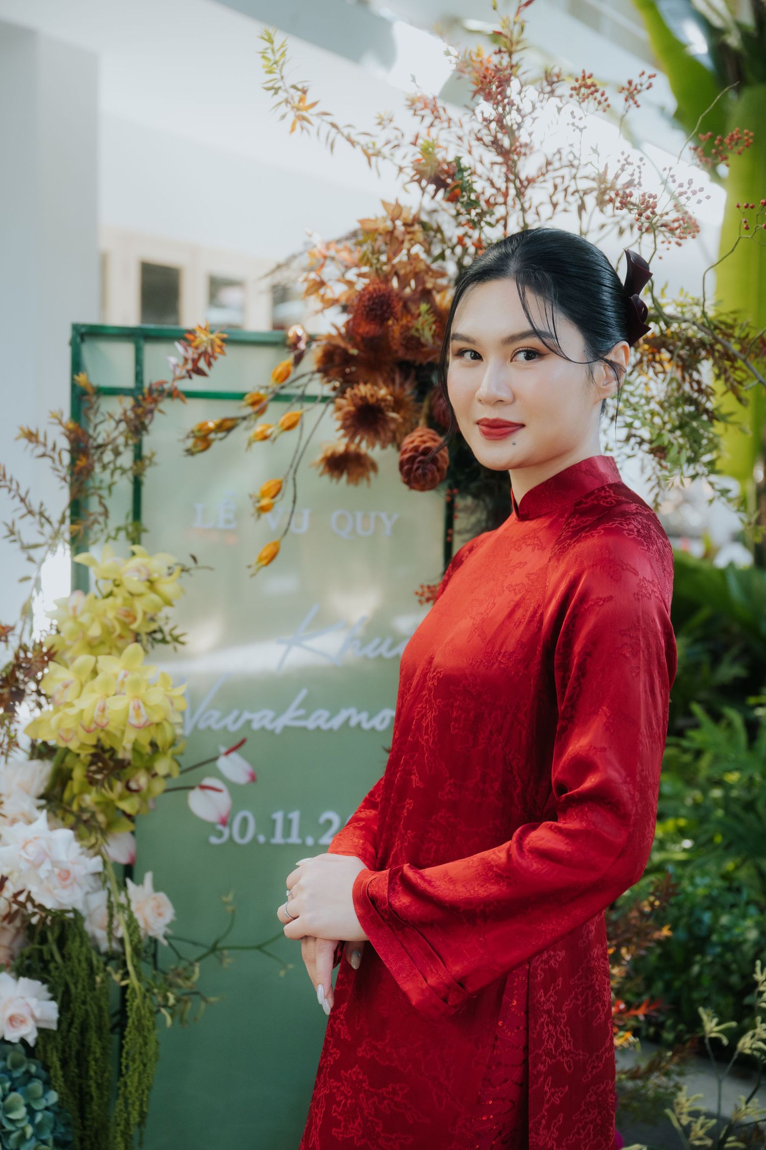 Young woman in red ao dai dress poses elegantly at a floral wedding backdrop with greenery and blooms.
