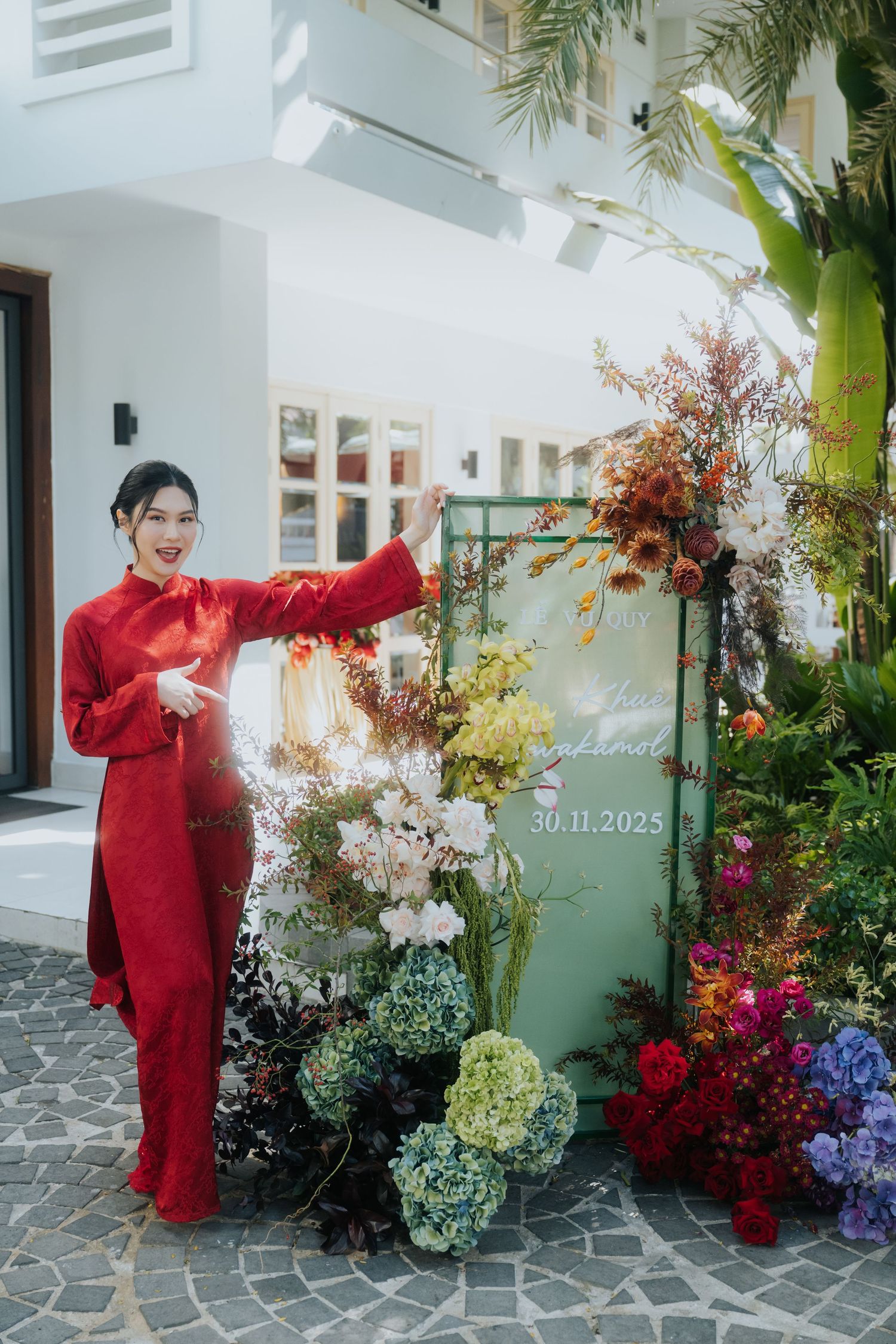A woman in a red ao dai dress poses beside a floral wedding display dated 30.11.2025.