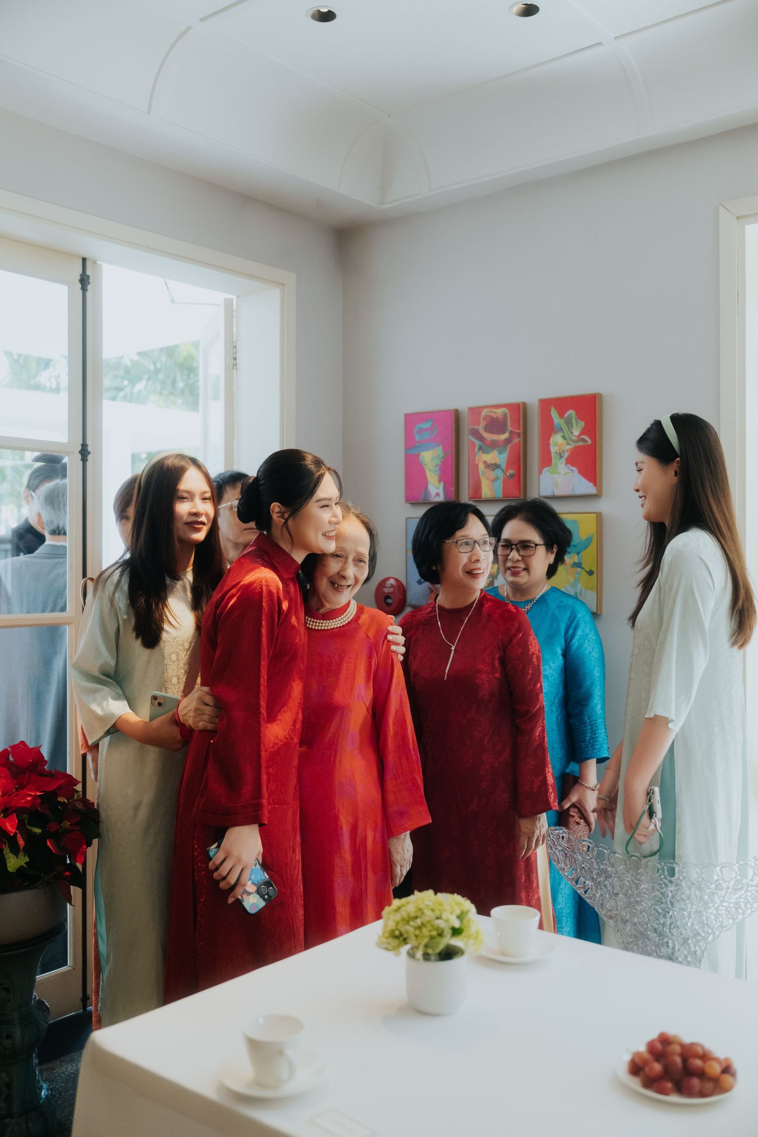 Group of Asian women in colorful traditional ao dai dresses gathered indoors during a festive celebration.
