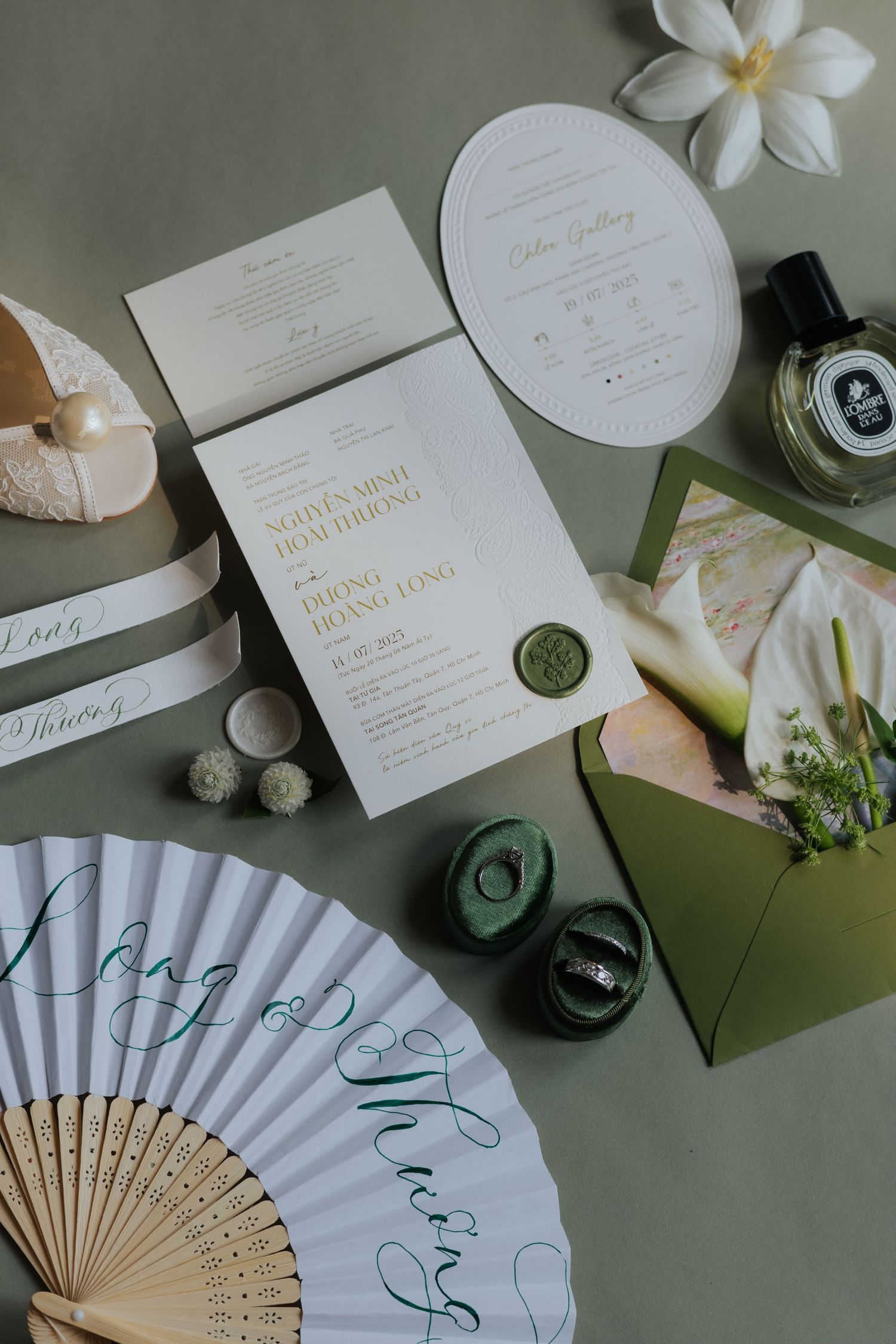 Elegant wedding stationery flatlay with gold foil invitations, wax seal, paper fans, and green envelope on sage background.