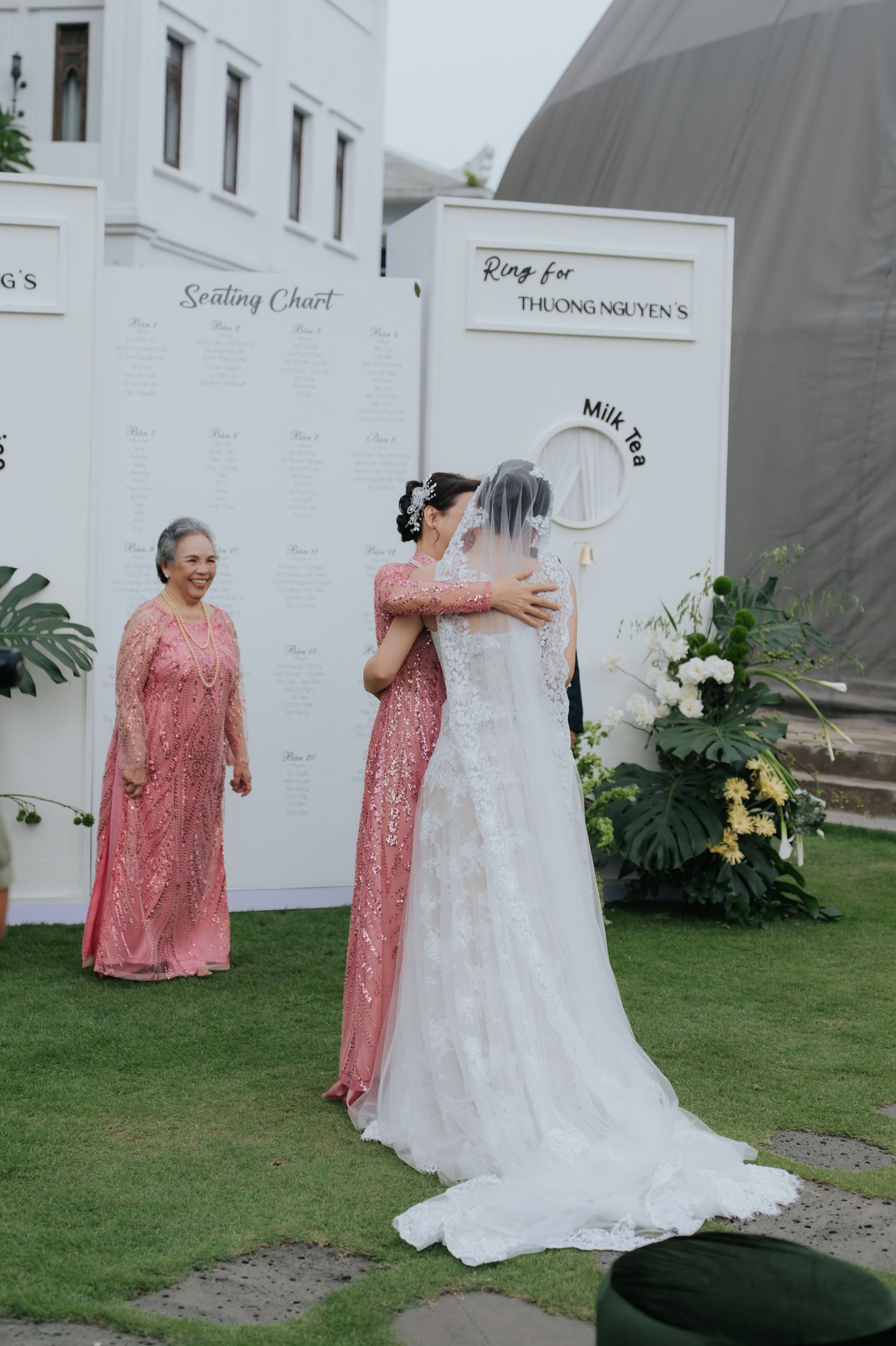 Bride in white gown embraces guest in pink dress at outdoor wedding with seating chart backdrop.