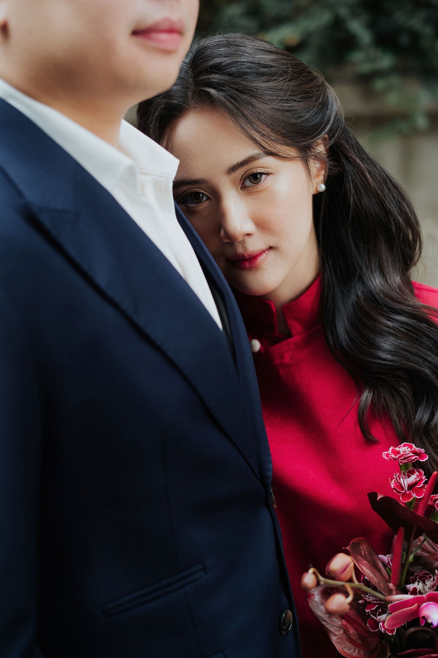 A woman in a red dress holds pink flowers, gazing at a man in a navy suit during a romantic couple photoshoot.