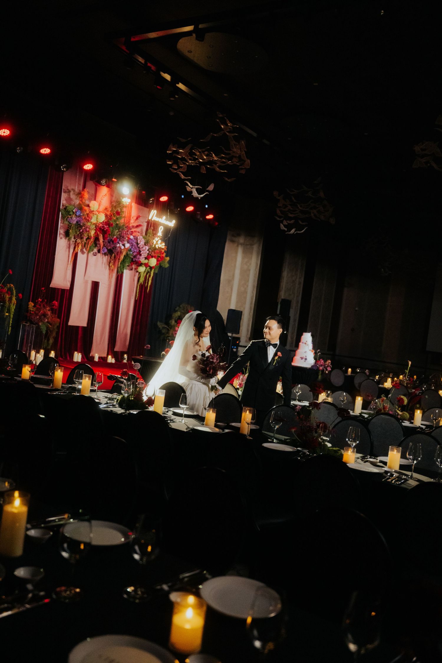 Elegant wedding reception with candlelit black tables, floral arrangements, and couple at head table in dimly lit venue.