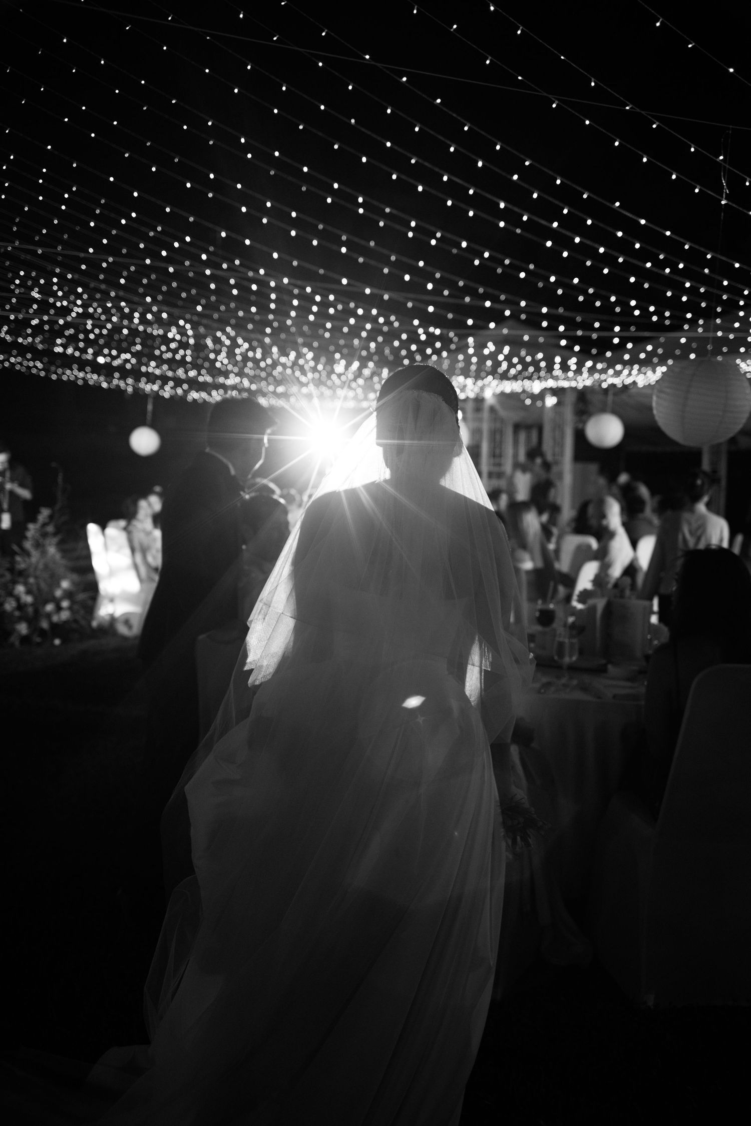 Black and white photo of bride and groom silhouetted against sparkling fairy lights at elegant wedding reception.