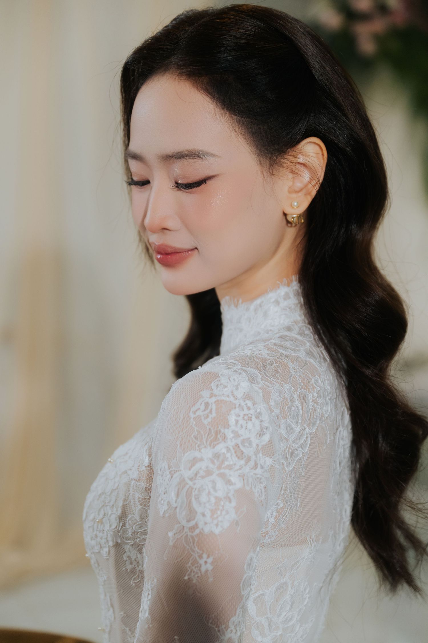 Elegant woman in white lace high-neck dress with gold earrings, eyes closed, dark wavy hair styled to side.