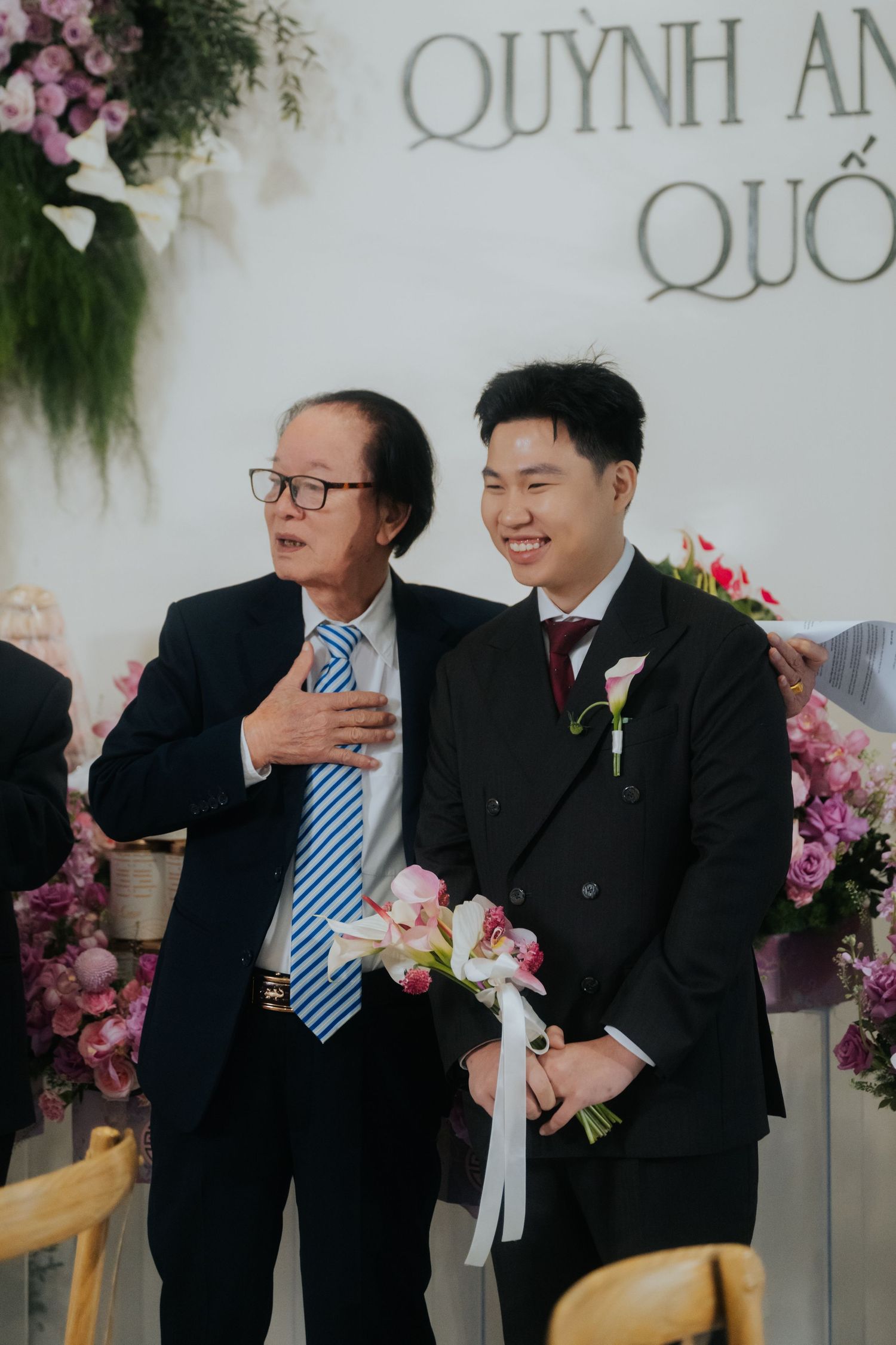 Two men in suits smile at a wedding ceremony with floral decorations and the names Quynh An and Quoc visible.