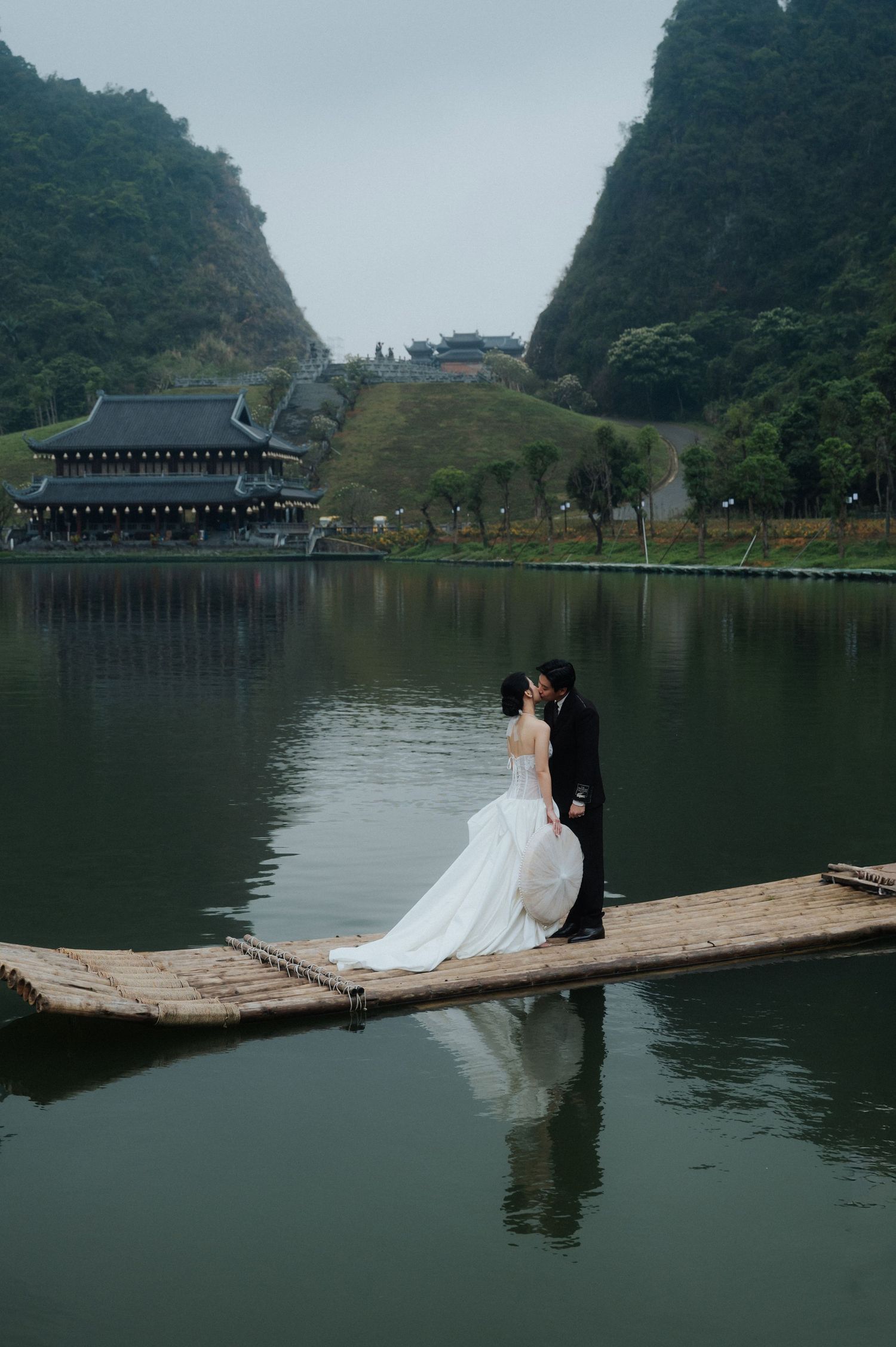 Bride and groom kissing on a bamboo raft over a calm lake with karst mountains and a temple in Vietnam.
