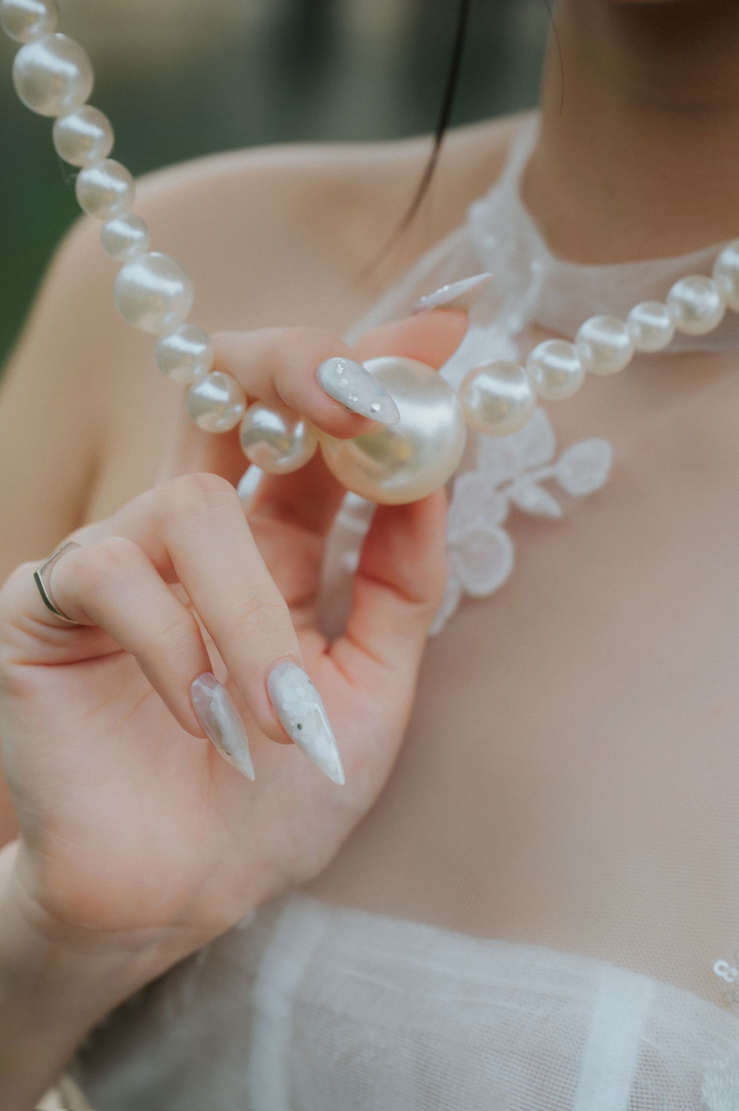 Woman with long white nails wearing a sheer white dress and elegant baroque pearl necklace, holding the pendant delicately.