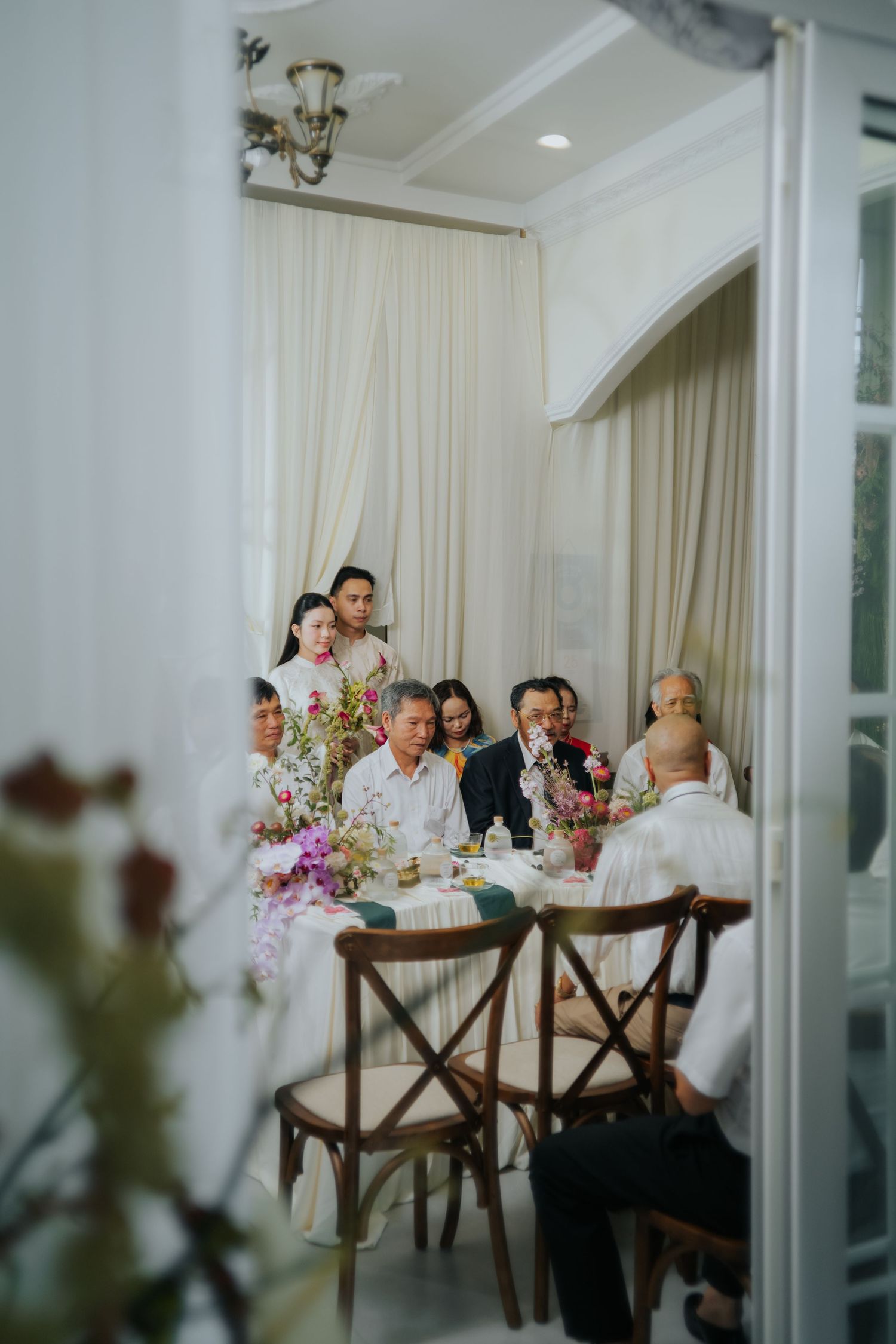 Elegant family gathering around a floral-decorated dining table in a white-draped room with wooden cross-back chairs.