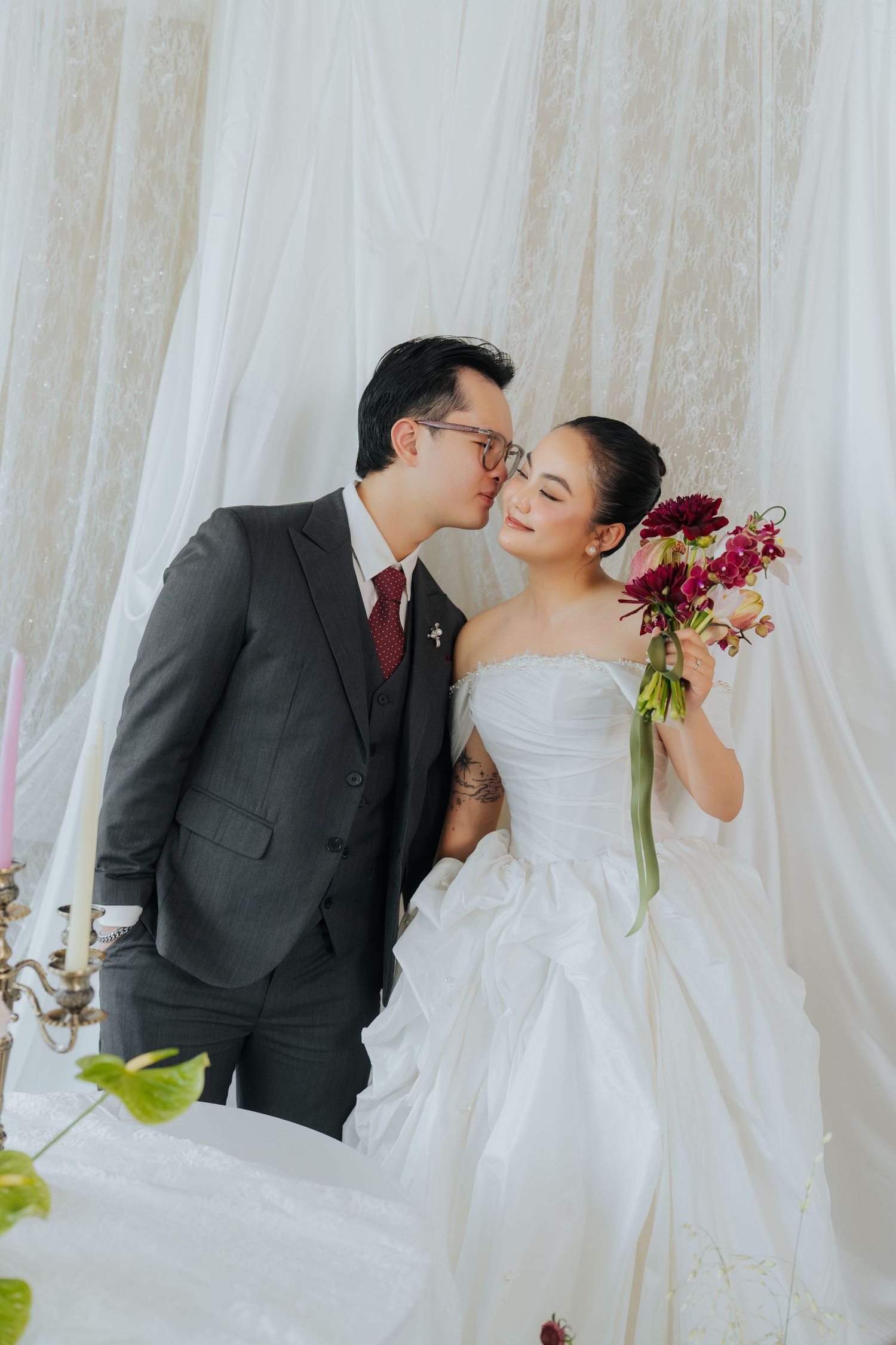 Groom in gray suit kisses bride in white strapless gown holding burgundy flowers against draped white backdrop.