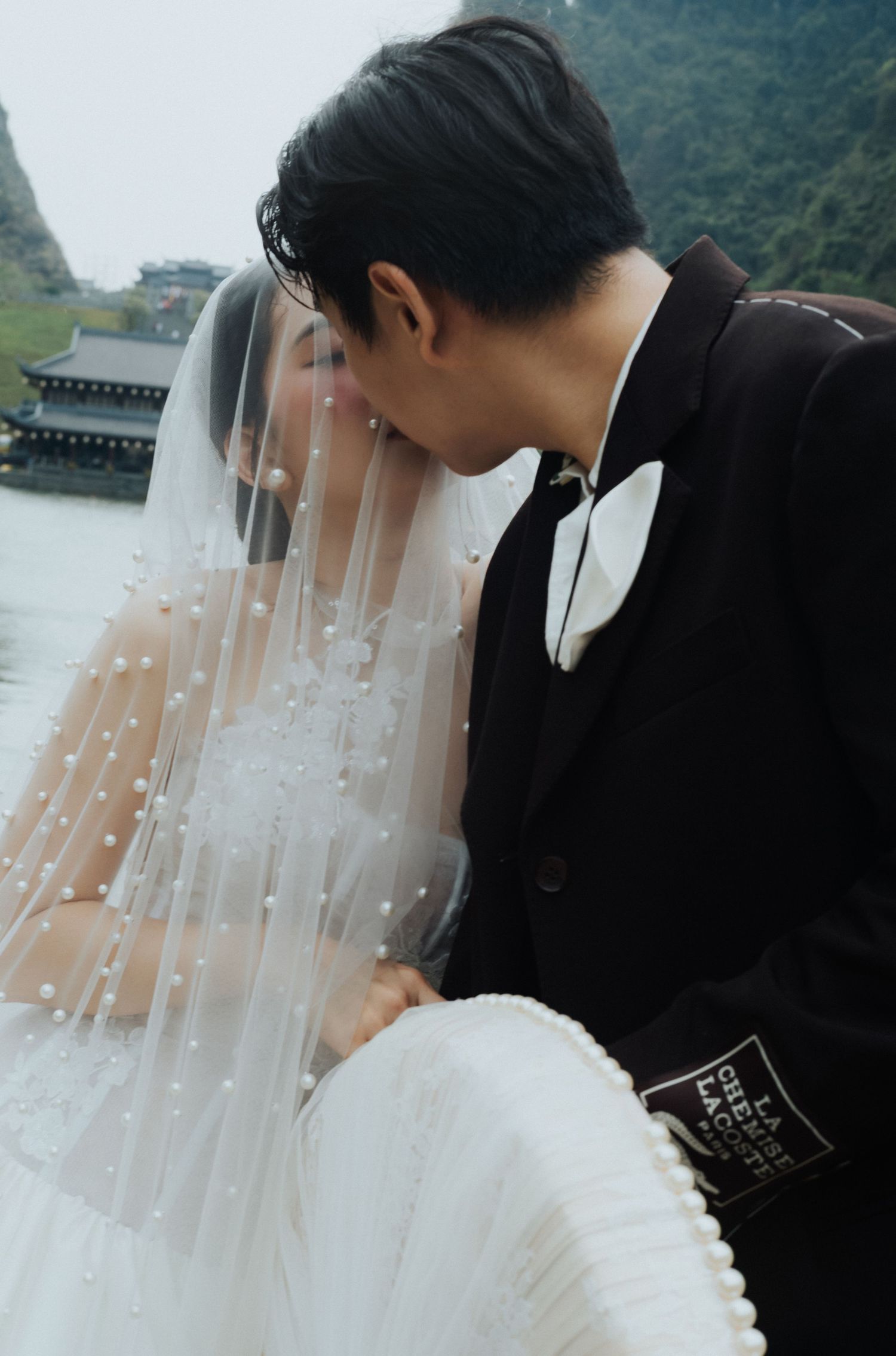 A couple shares a romantic kiss in wedding attire, with a pearl-adorned veil and black suit, set against a scenic waterway.
