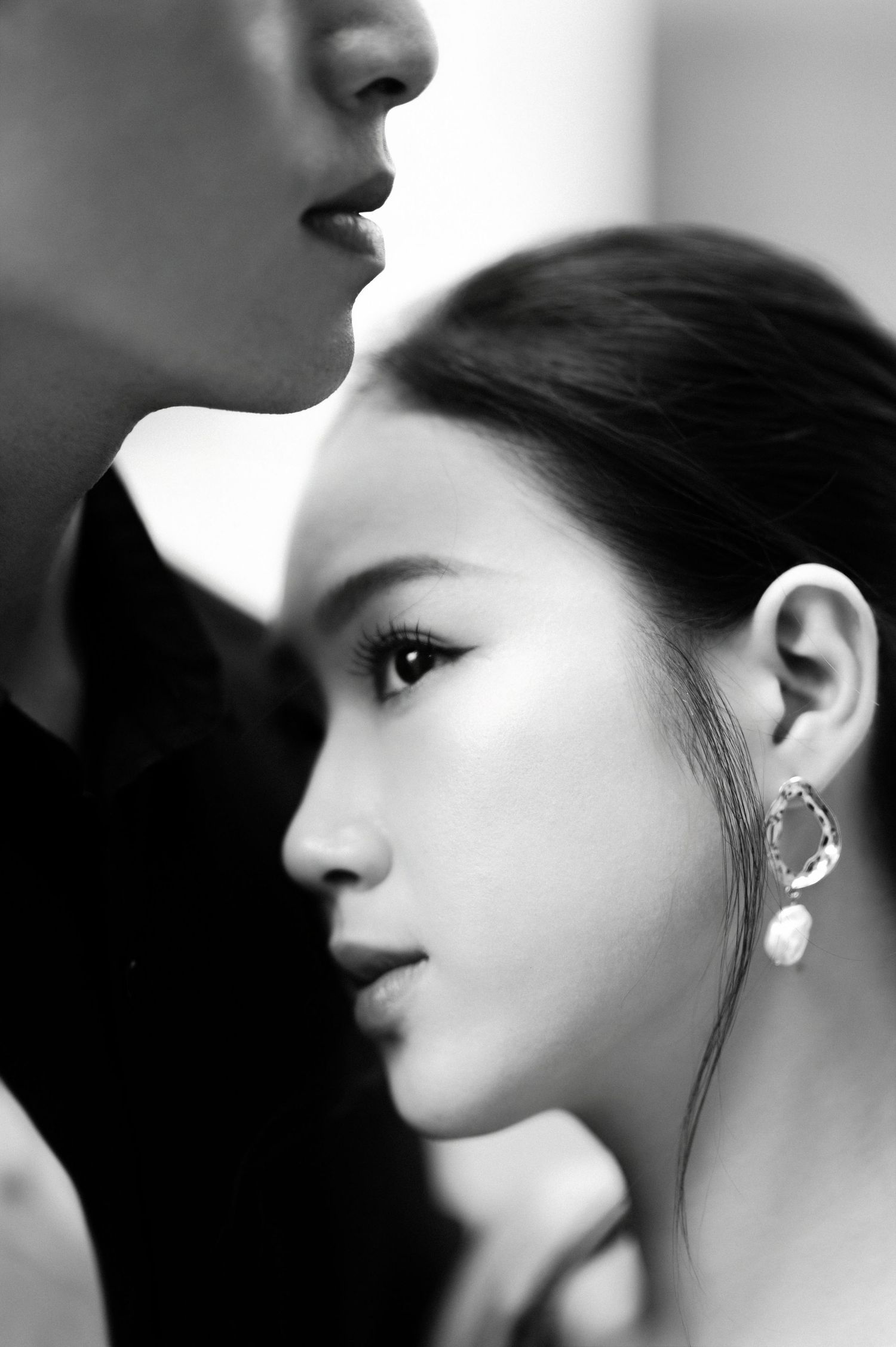 Black and white portrait of an elegant Asian woman in profile wearing pearl earrings, with a man close behind her.