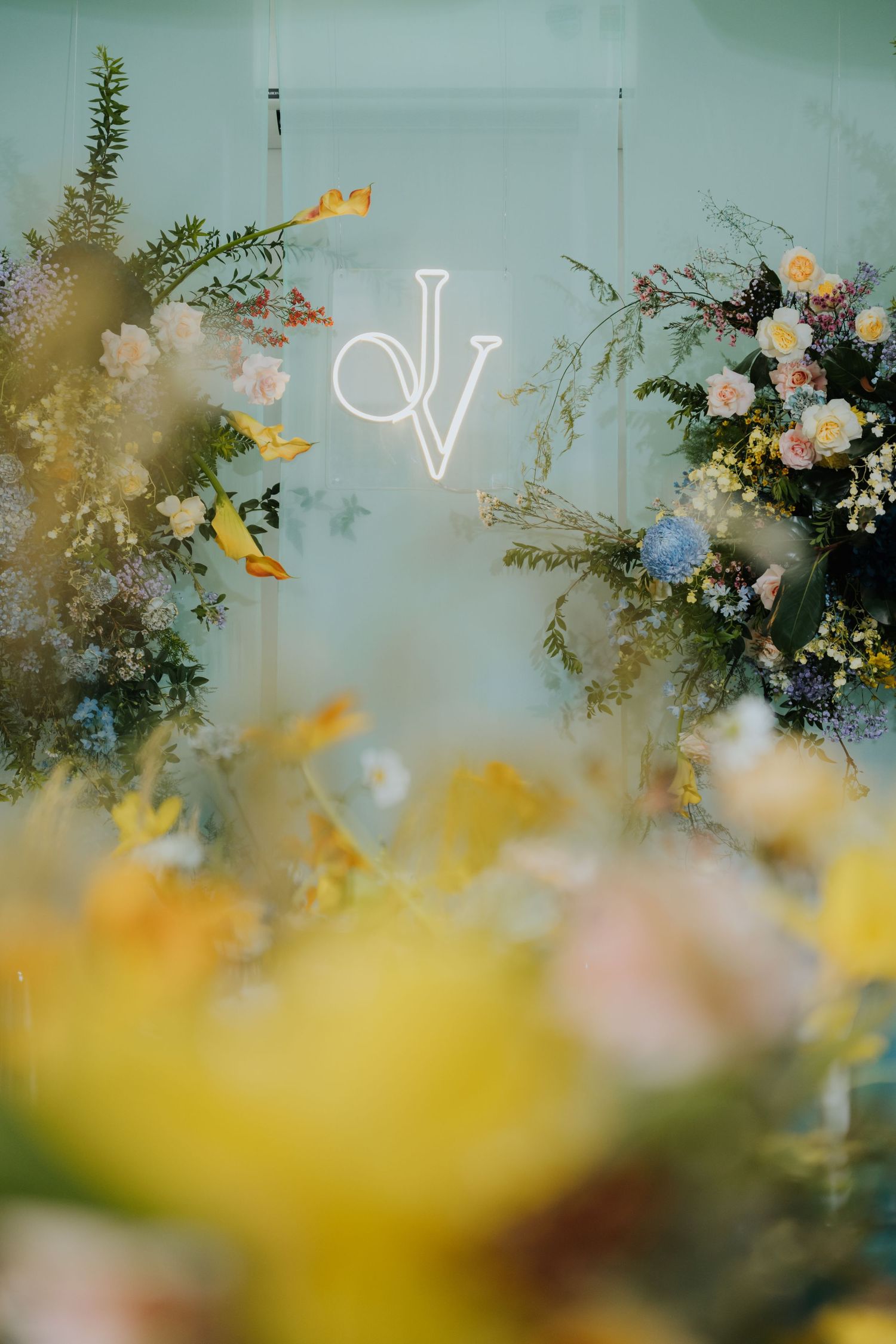 Elegant wedding backdrop with neon monogram JV sign surrounded by lush floral arrangements in yellow, blue, and pink blooms.