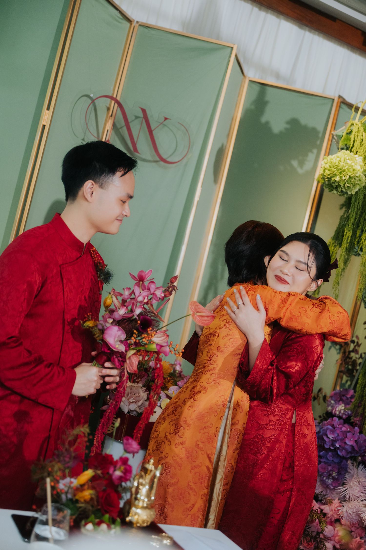 Two people in red traditional Vietnamese ao dai embrace at a wedding ceremony with floral decorations.