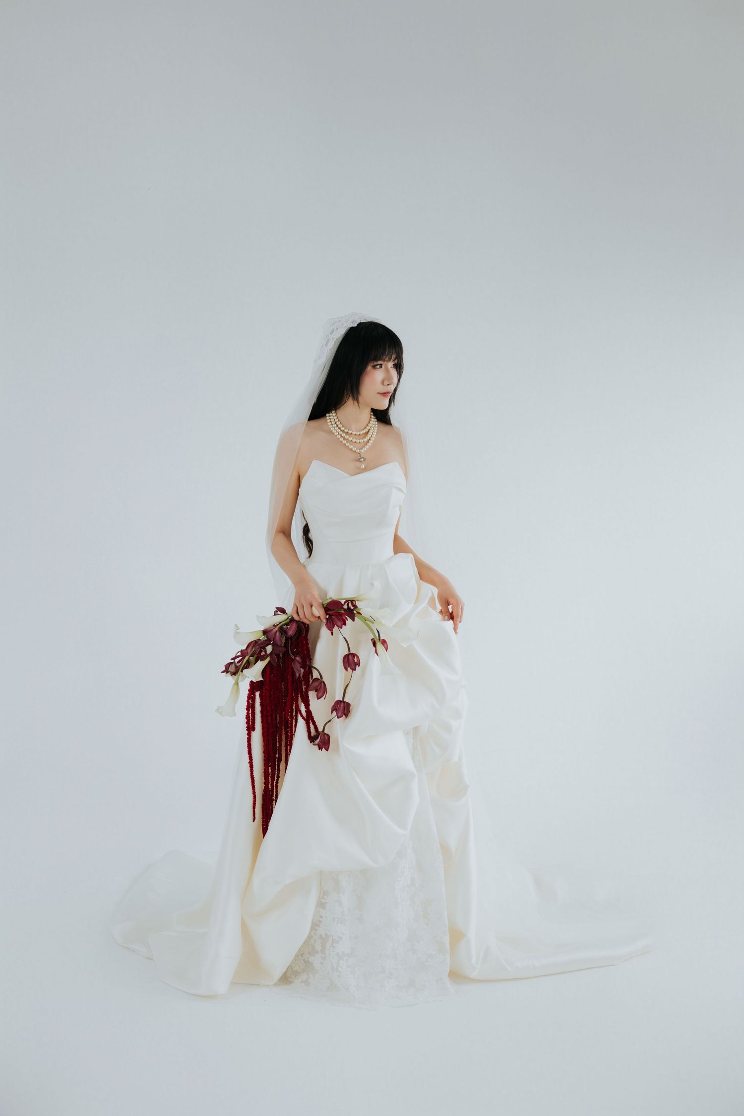 Elegant bride in strapless white ballgown with train holding a cascading burgundy floral bouquet against white background.