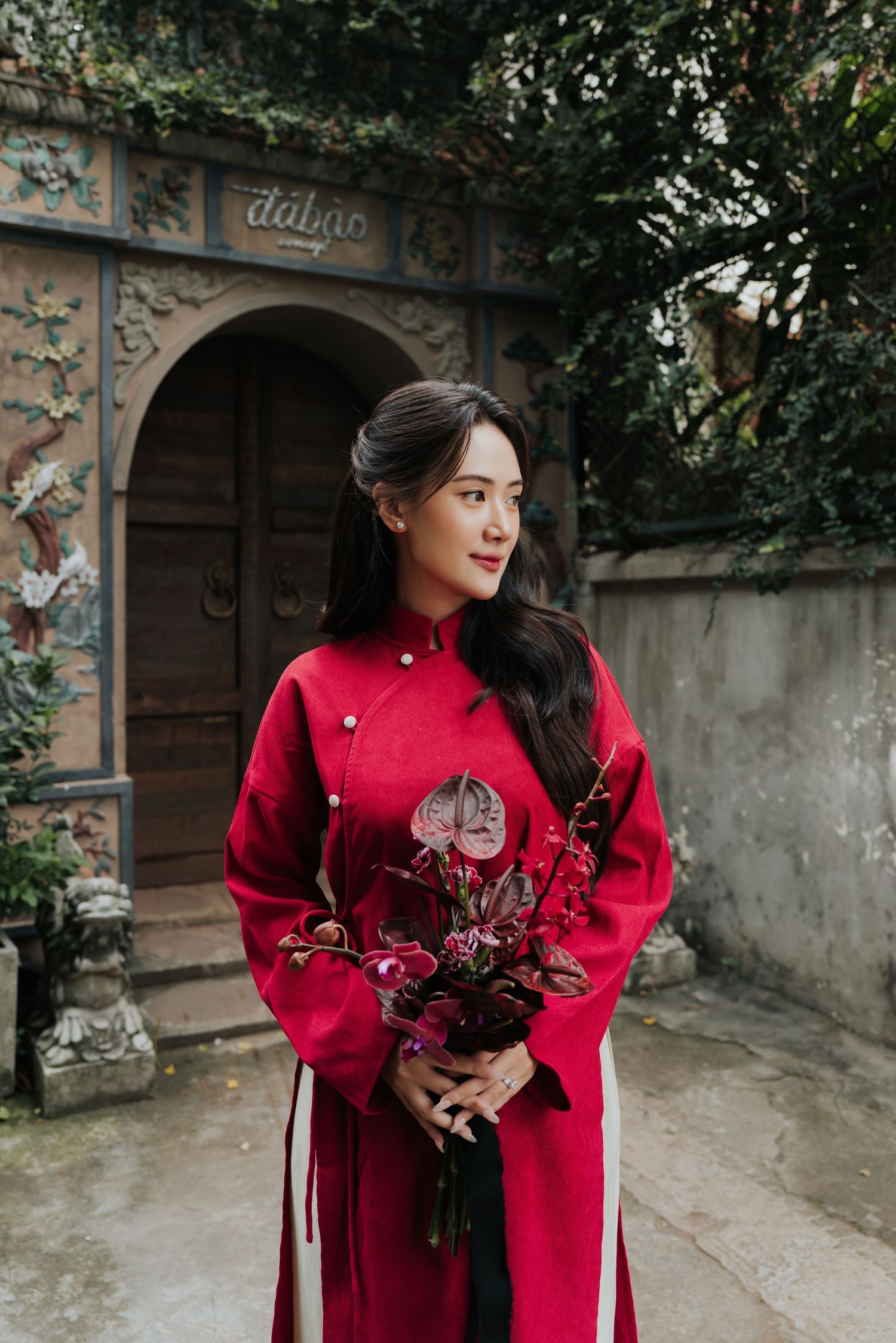 Woman in red ao dai holding burgundy floral bouquet stands before ornate stone archway in traditional Vietnamese setting.