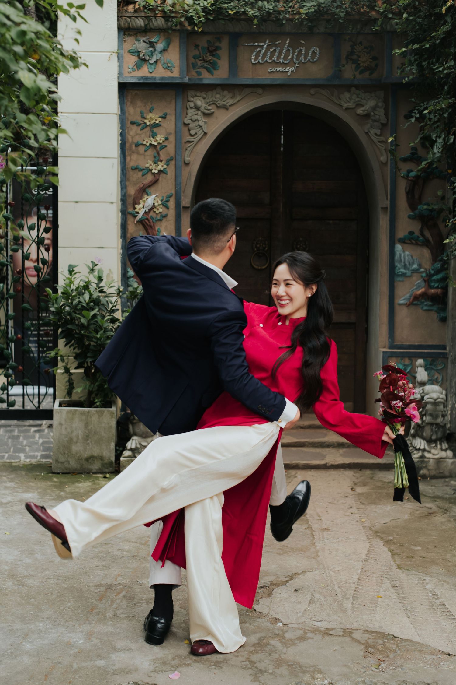 Couple in traditional Vietnamese attire dancing joyfully outdoors, woman in red ao dai holding flowers, man in dark suit.