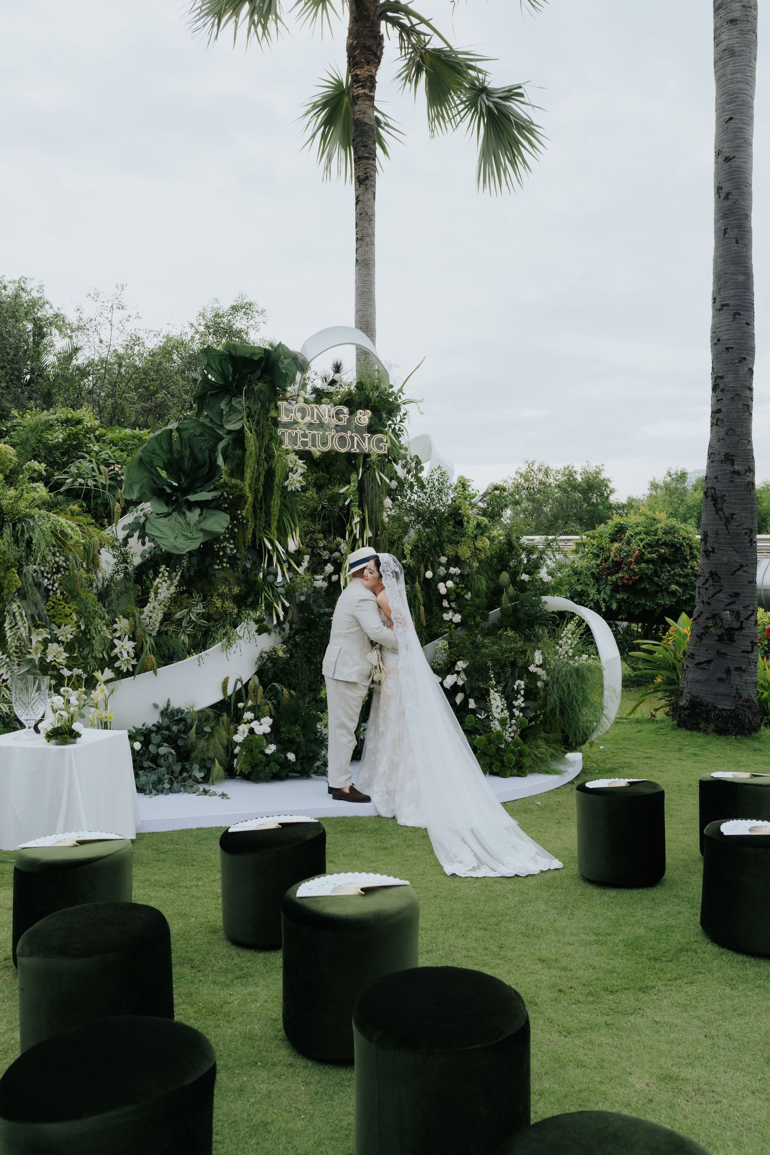Couple shares a kiss at an outdoor garden wedding ceremony with lush greenery backdrop and elegant white floral arrangements.