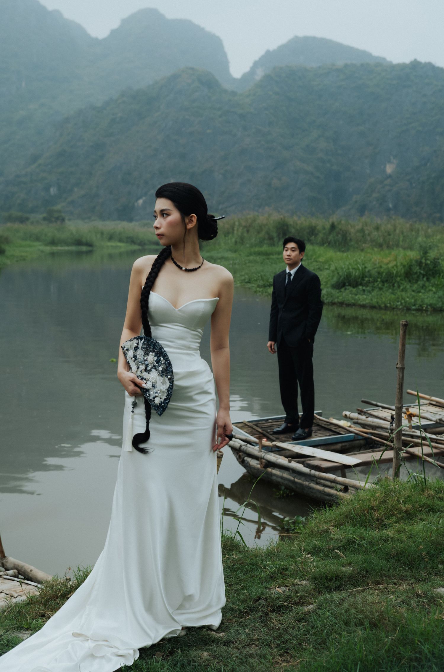Bride in white strapless gown holds fan by river, groom in black suit stands on boat, misty mountains backdrop.