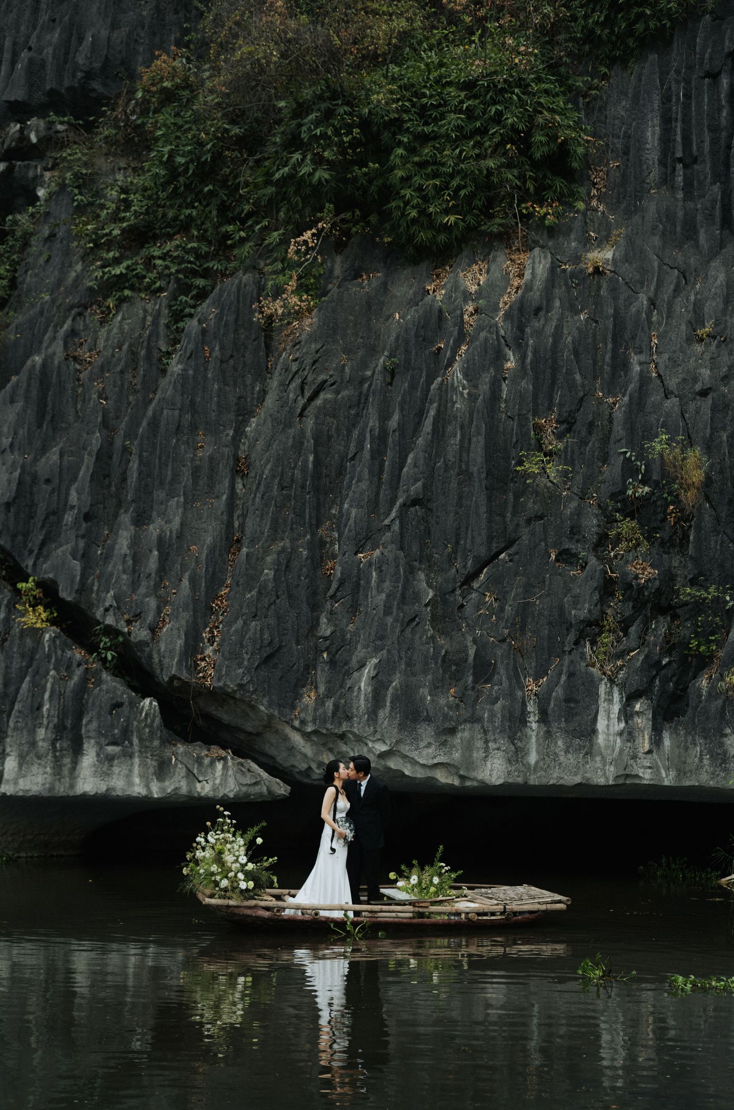 Couple shares a romantic kiss on a flower-adorned boat, reflected in calm water before dramatic dark cliffs.