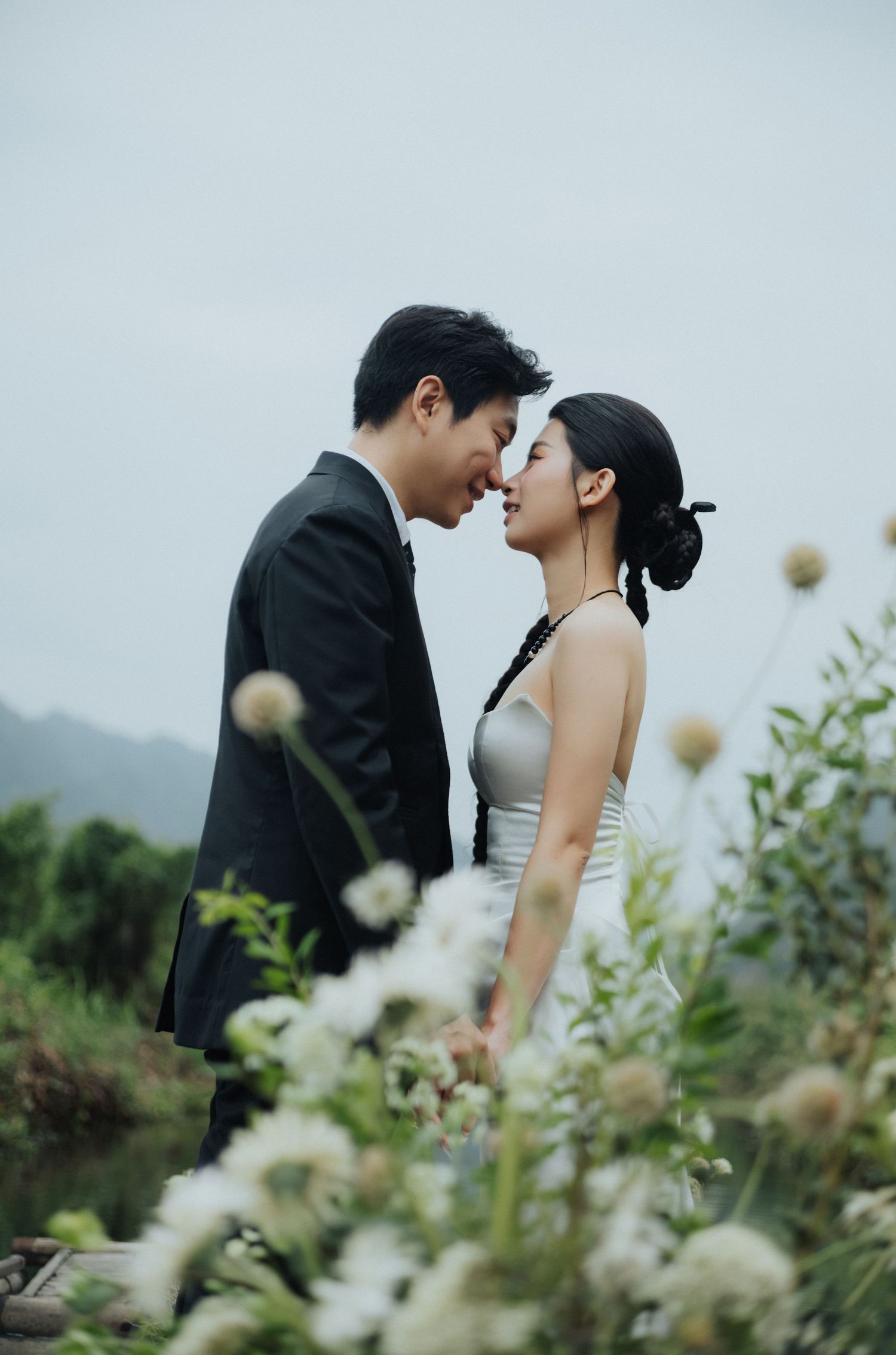 A couple in wedding attire shares an intimate moment surrounded by wildflowers with misty mountains in the background.