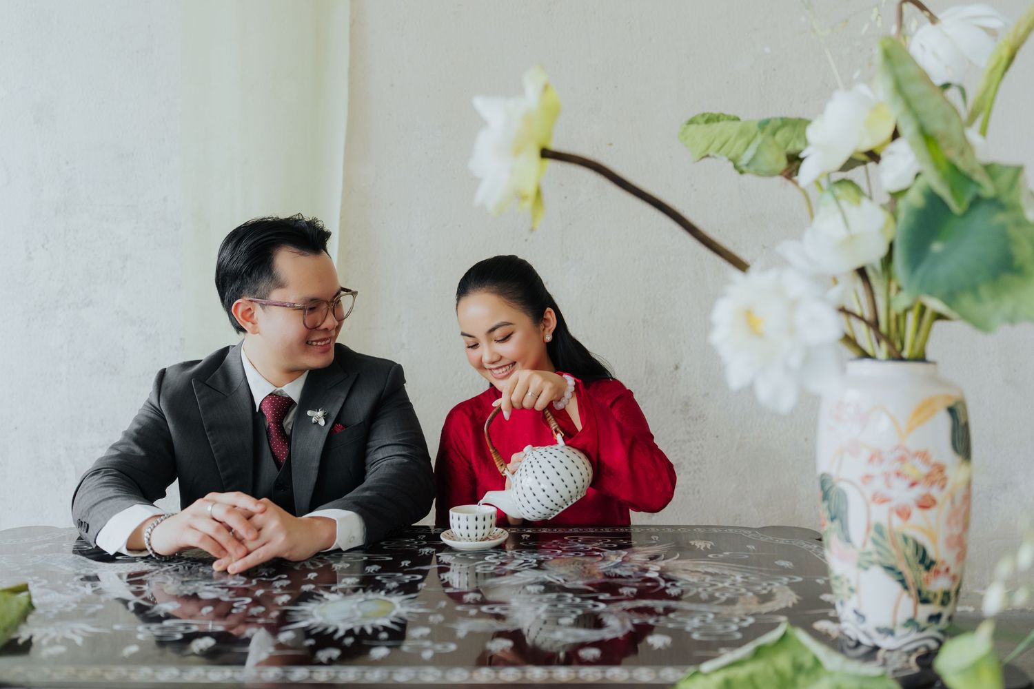 A couple shares a romantic tea ceremony moment, with a woman in red pouring tea while a man in a suit smiles warmly.