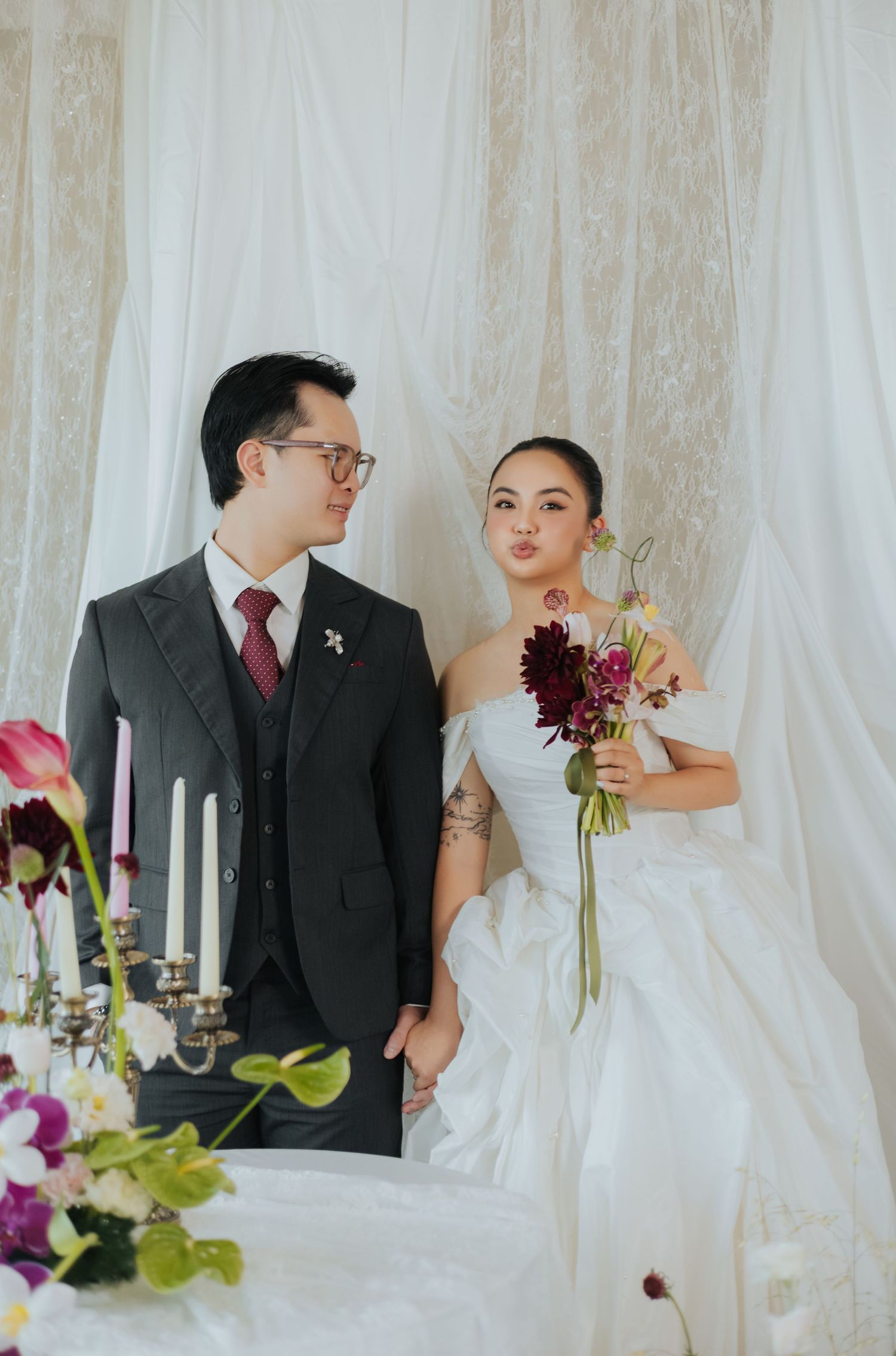 A couple in wedding attire poses by a floral table, the bride holding a bouquet and groom in a dark suit with red tie.