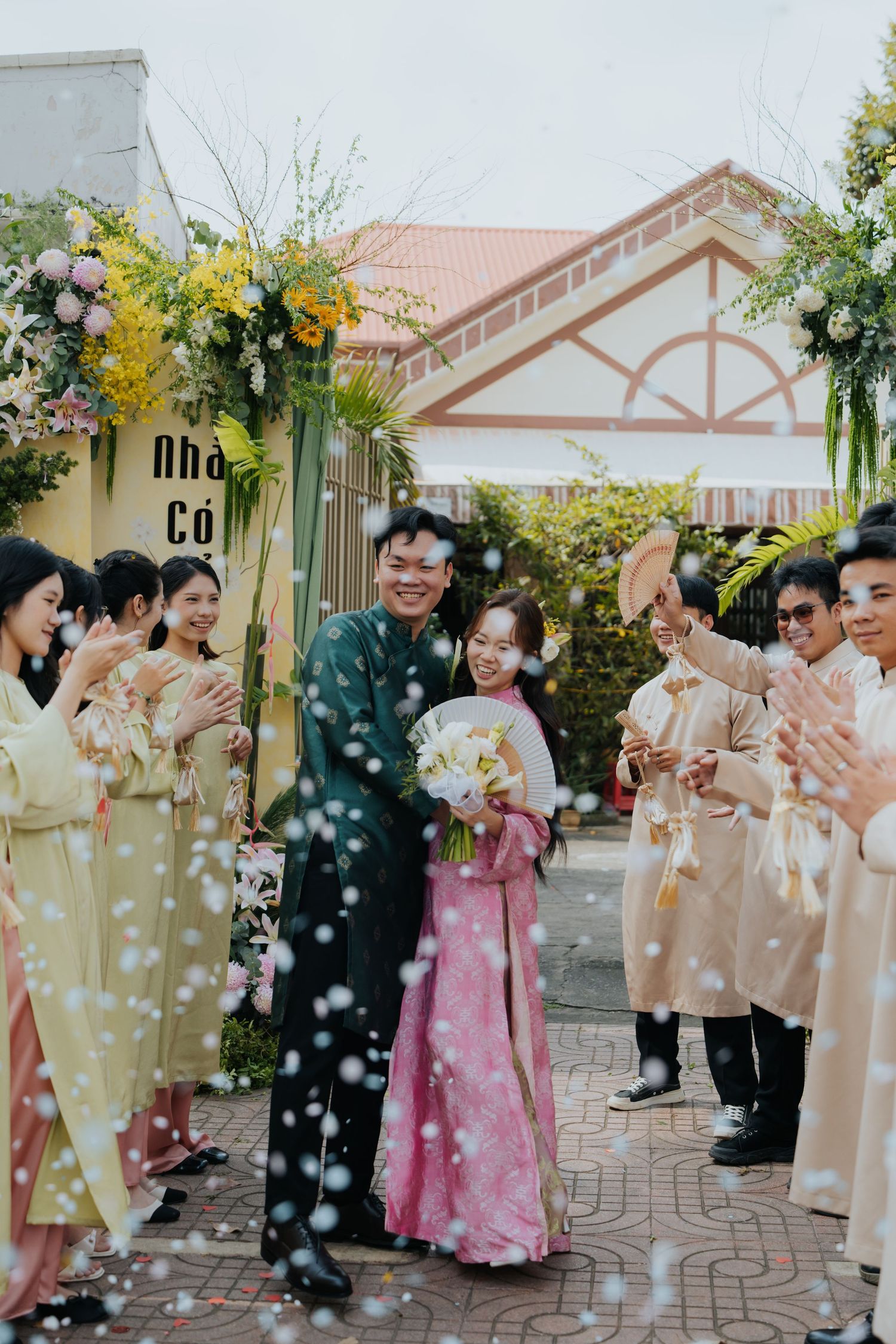 Vietnamese couple in traditional ao dai attire walks through confetti shower at outdoor wedding ceremony with floral arch.