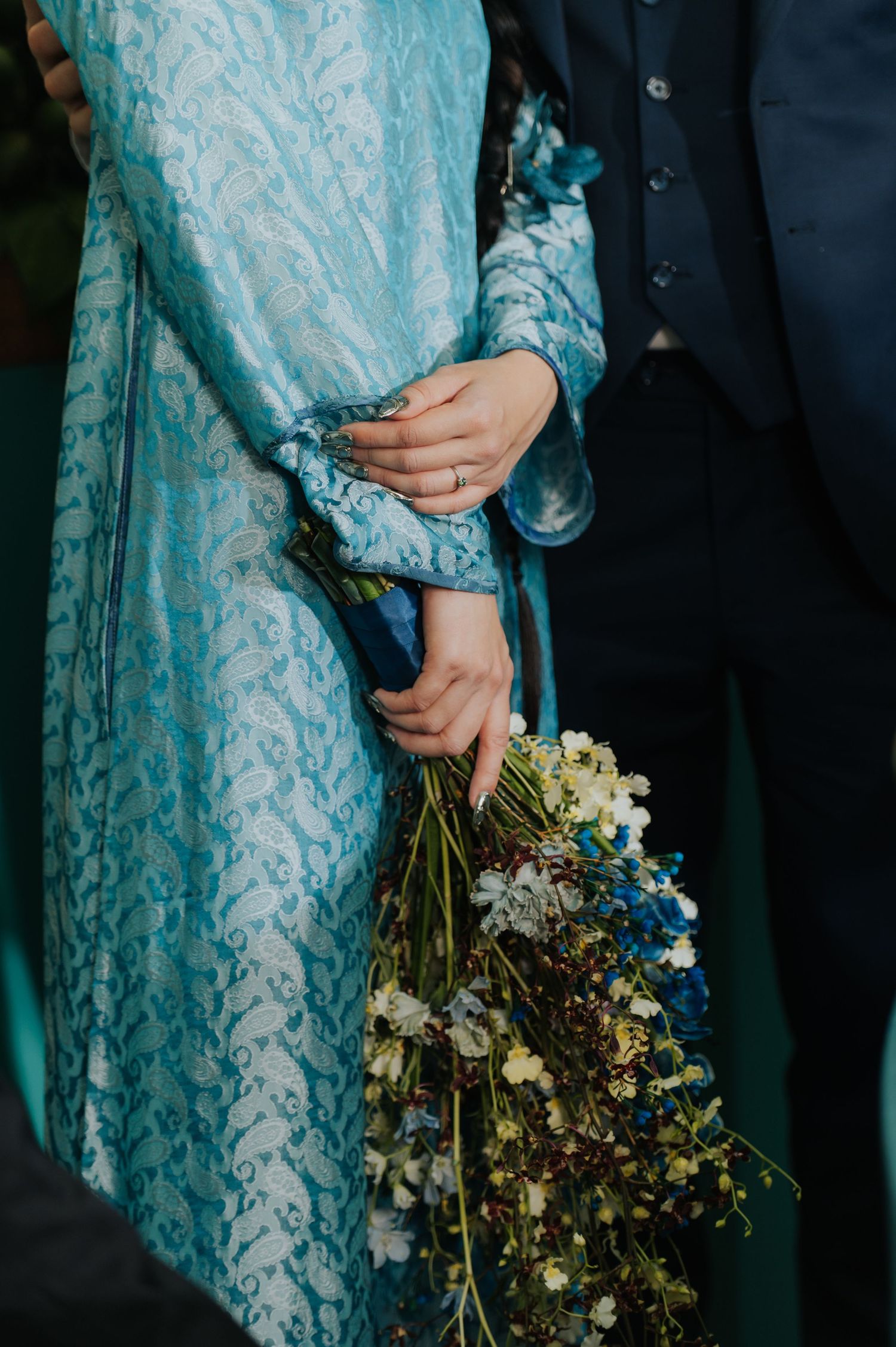 Bride in blue brocade dress holding wildflower bouquet beside groom in navy suit at wedding ceremony.