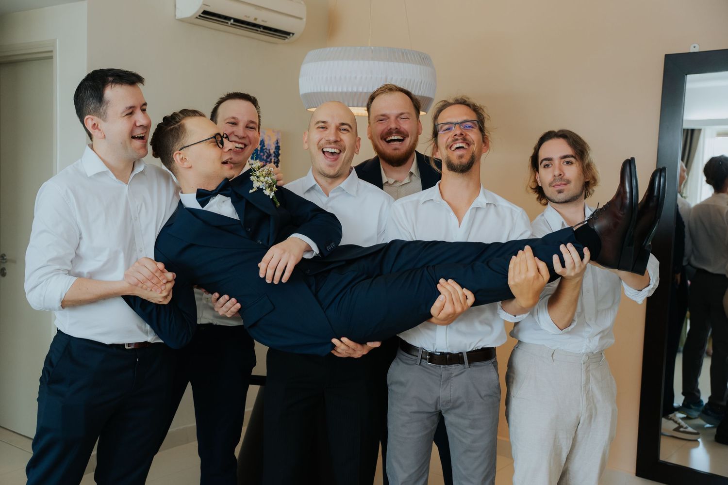 Groomsmen laughing and lifting the groom horizontally in a bright room during wedding preparations.