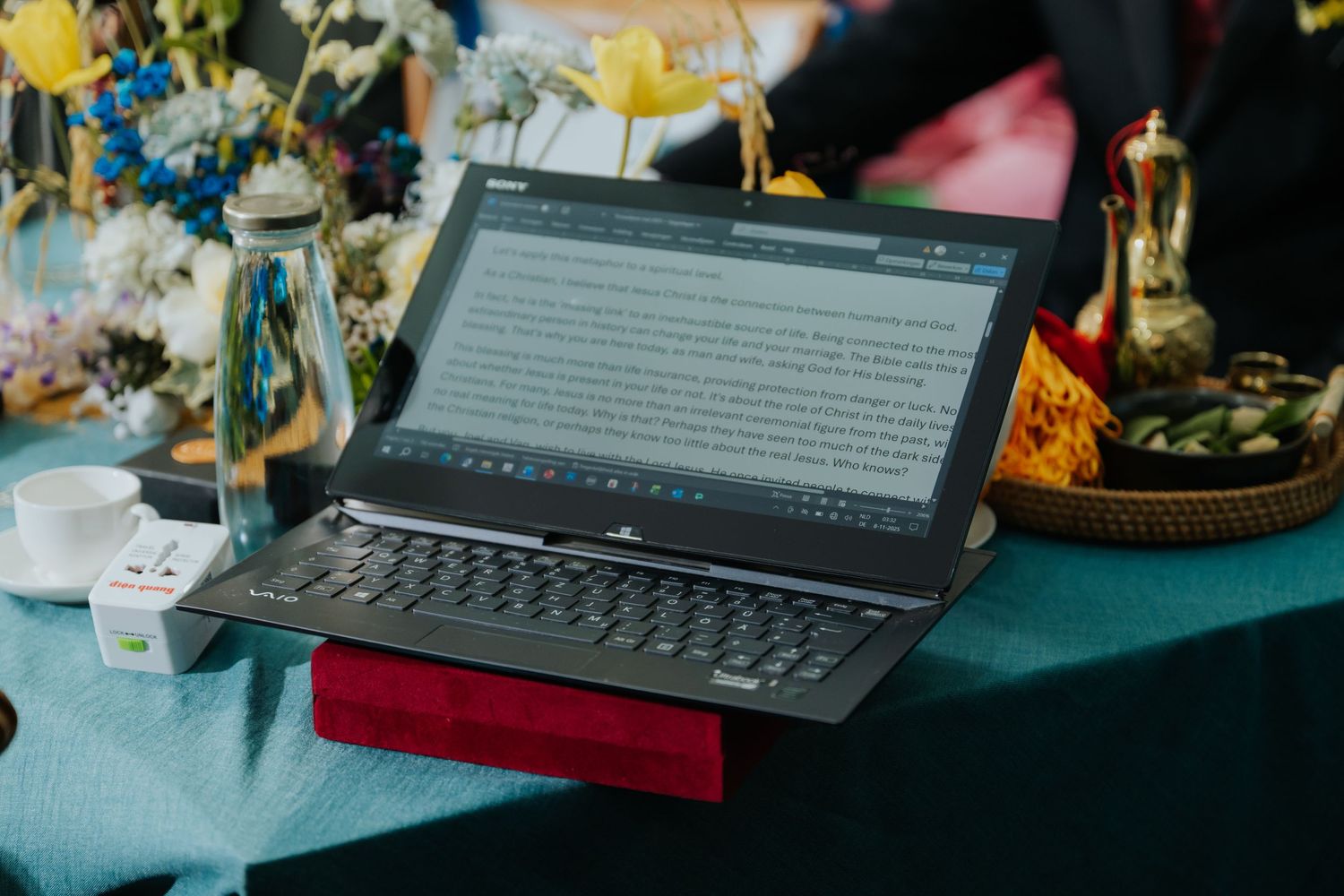 A laptop with a detachable keyboard displaying text, placed on a teal tablecloth with flowers and decorative items.