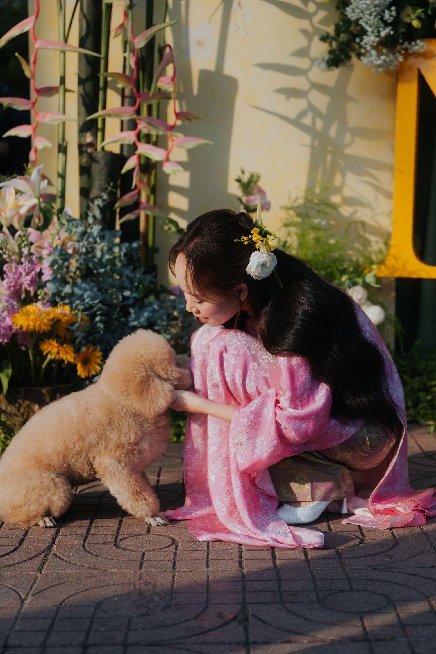 Young woman in pink kimono crouching to pet a fluffy cream poodle surrounded by colorful garden flowers.