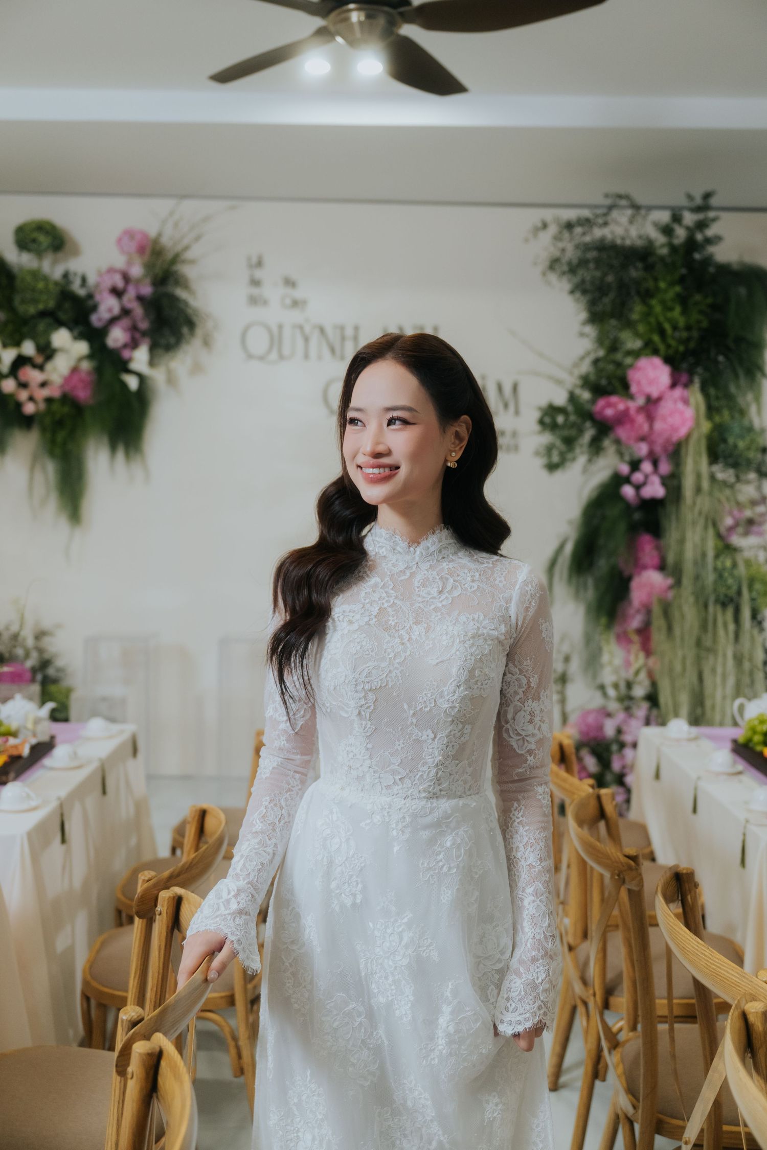 A bride in a white lace long-sleeve gown smiles in a floral-decorated wedding venue with wooden chairs.