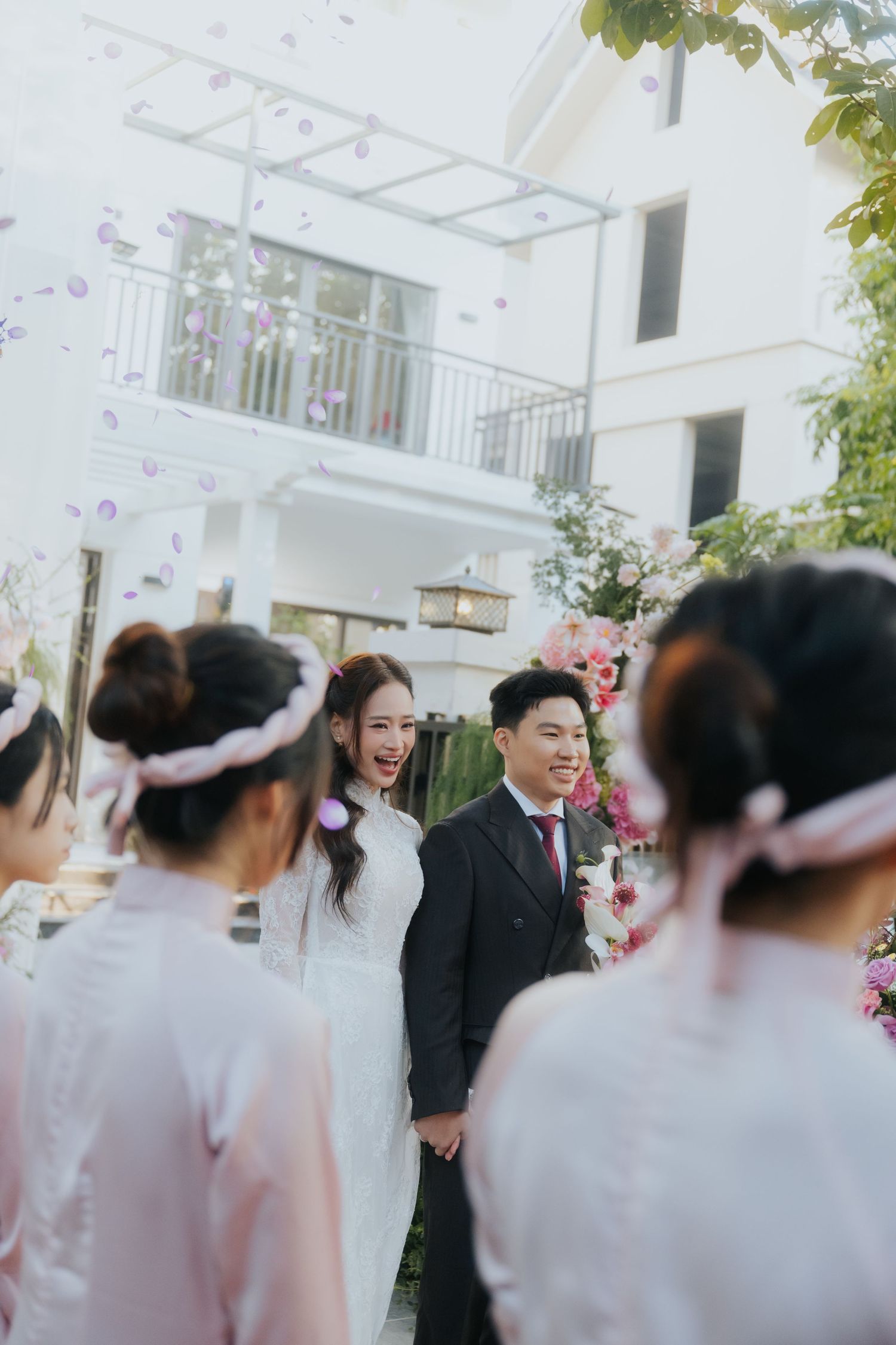A joyful bride and groom hold hands outdoors at a Vietnamese wedding ceremony with bridesmaids in pink ao dai.